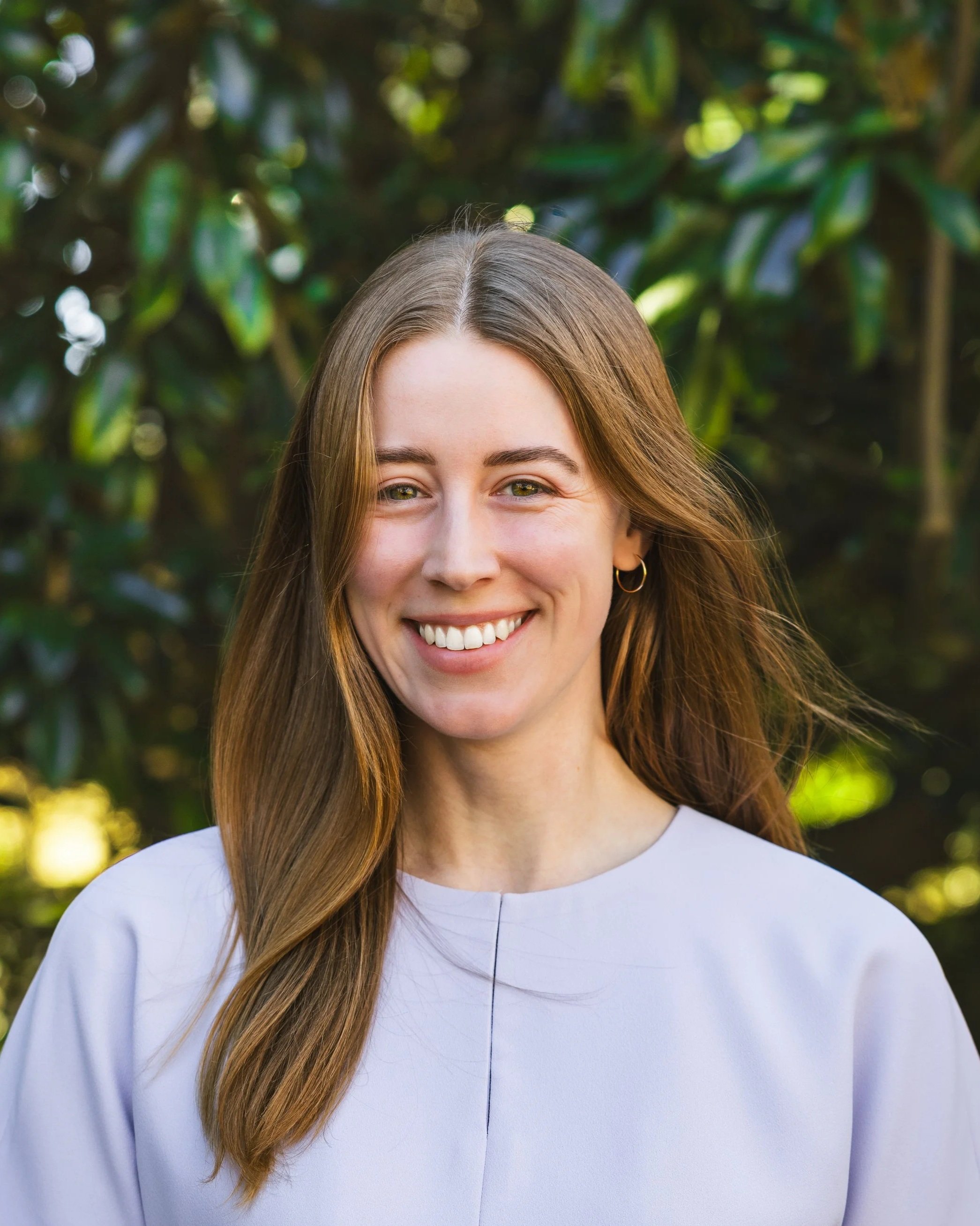 A woman with long brown hair smiling outdoors with green foliage in the background.