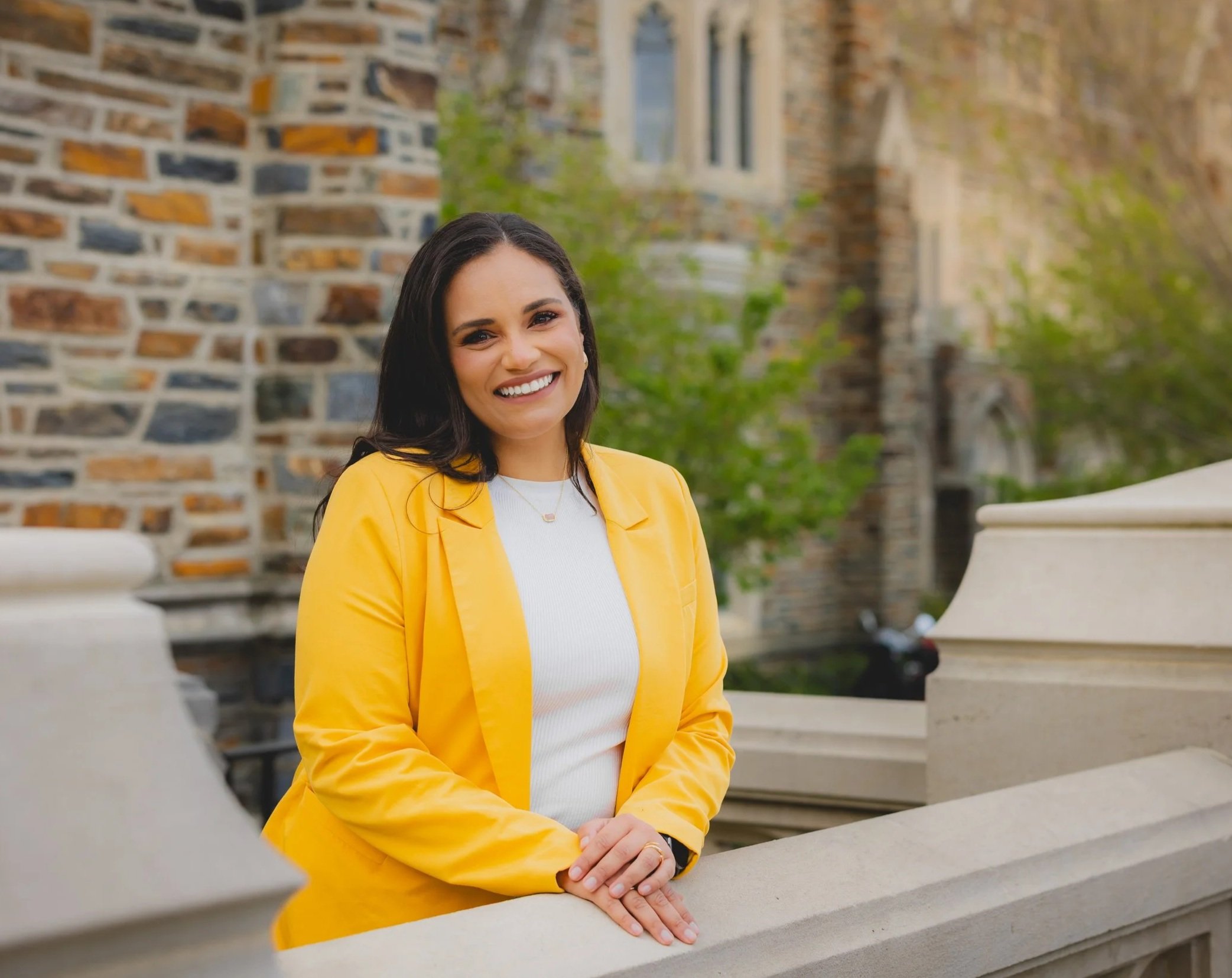 A woman with long dark hair, wearing a white top and yellow blazer, smiling outdoors in front of a stone building with greenery.