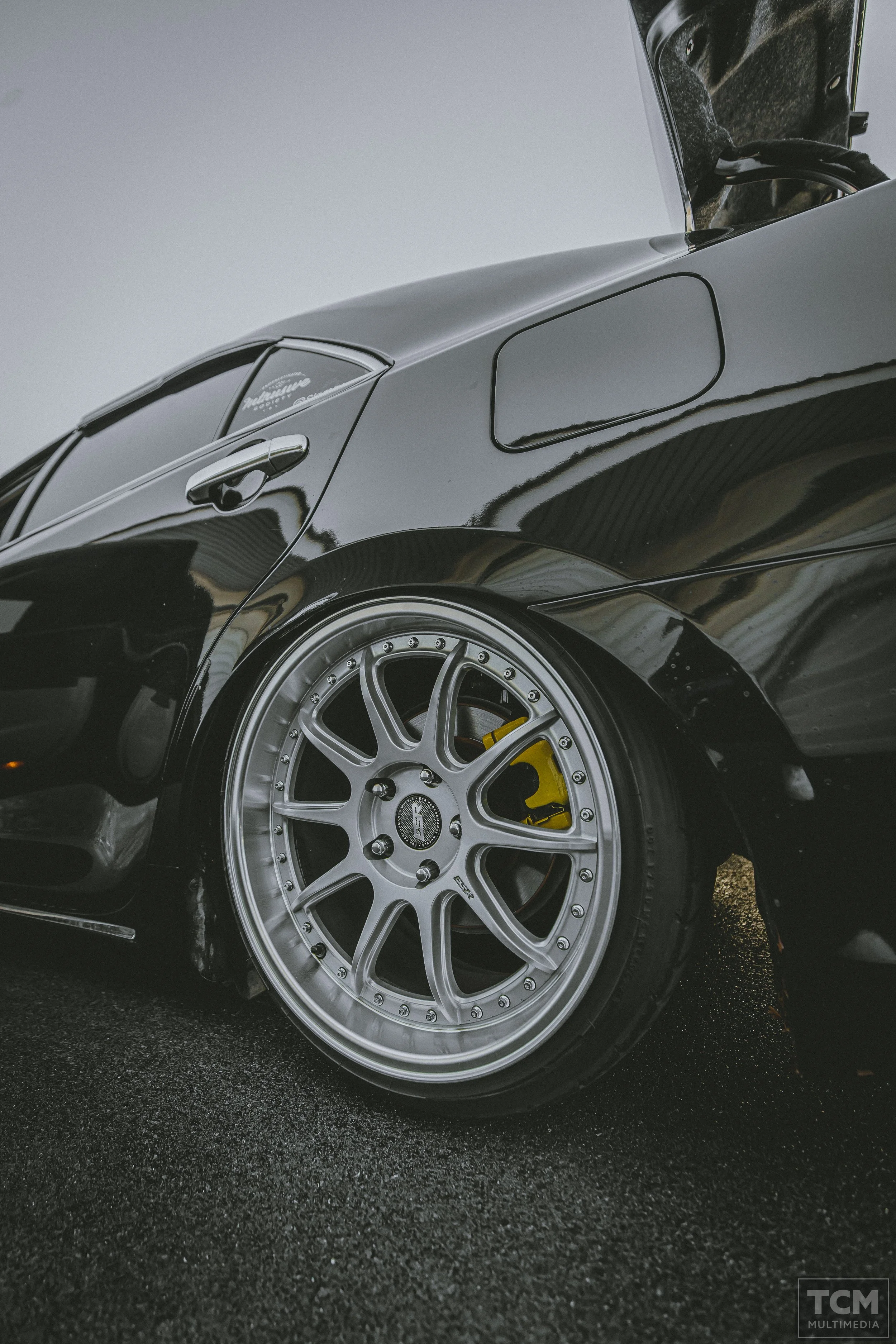 Close-up of a black sports car, showing the rear wheel with silver rims and yellow brake caliper.