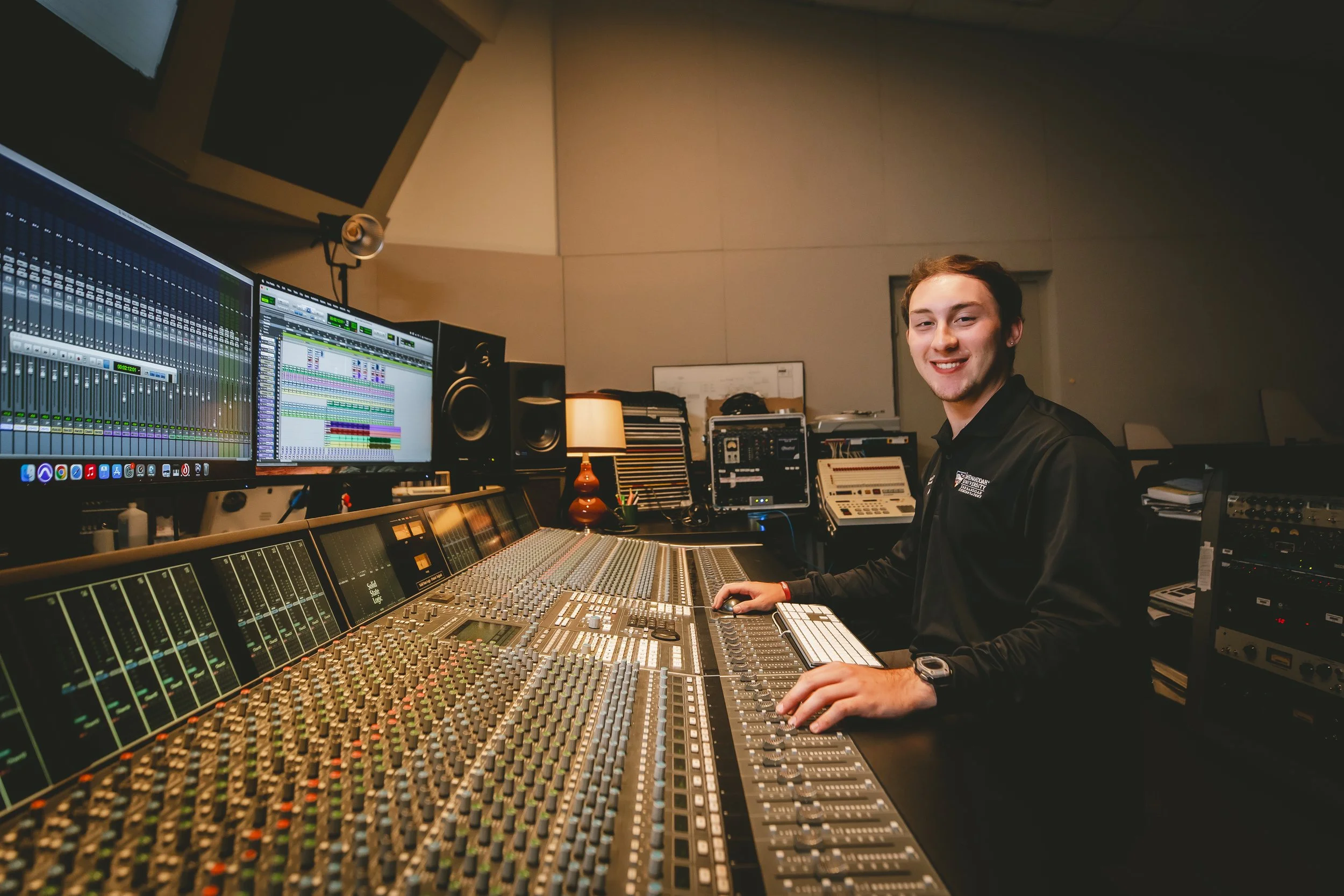 A young man smiling at a music mixing console in a recording studio with multiple monitors, speakers, and audio equipment.