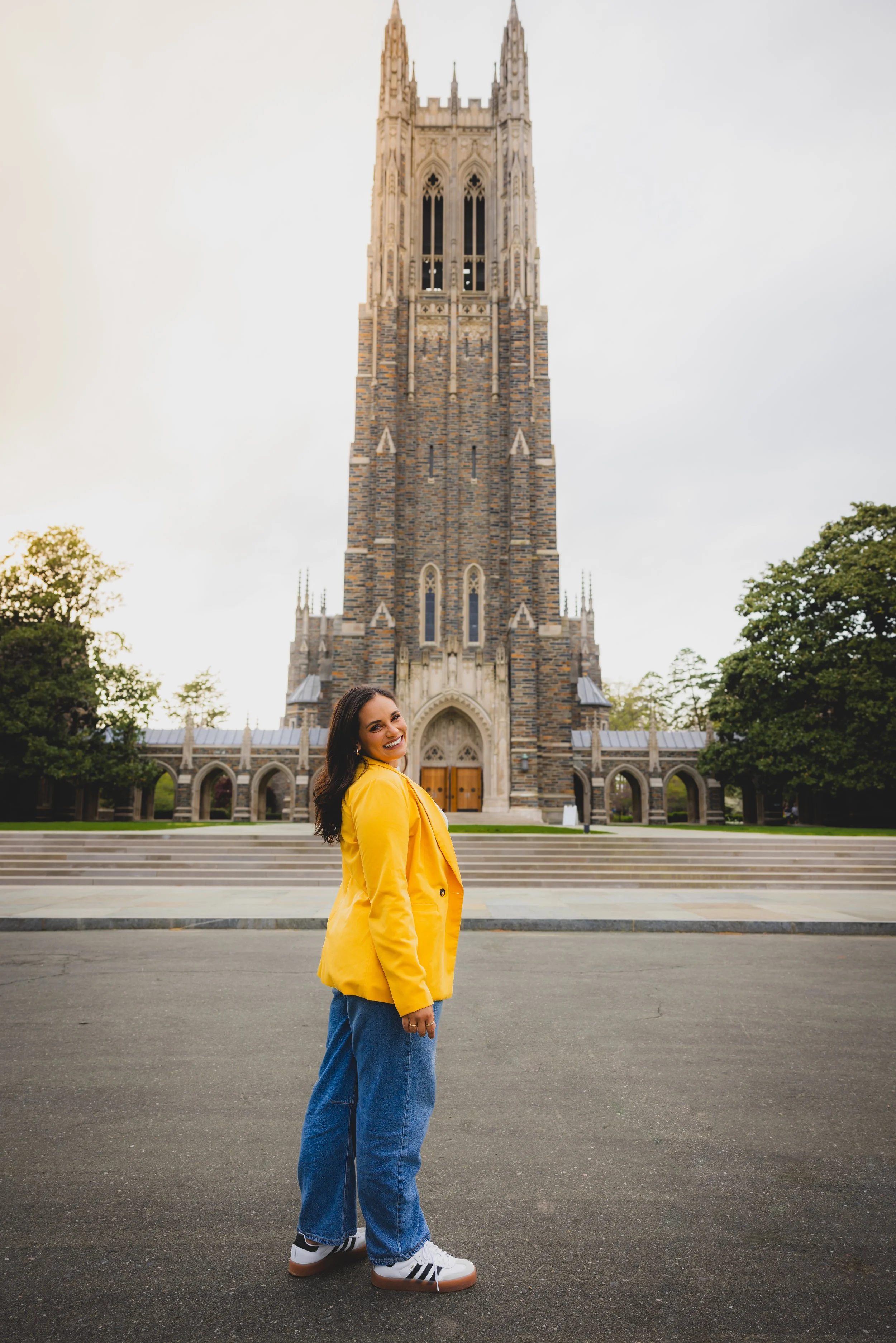 A woman in a yellow jacket and blue jeans standing on a paved area in front of a large Gothic-style church with a tall tower, surrounded by trees.