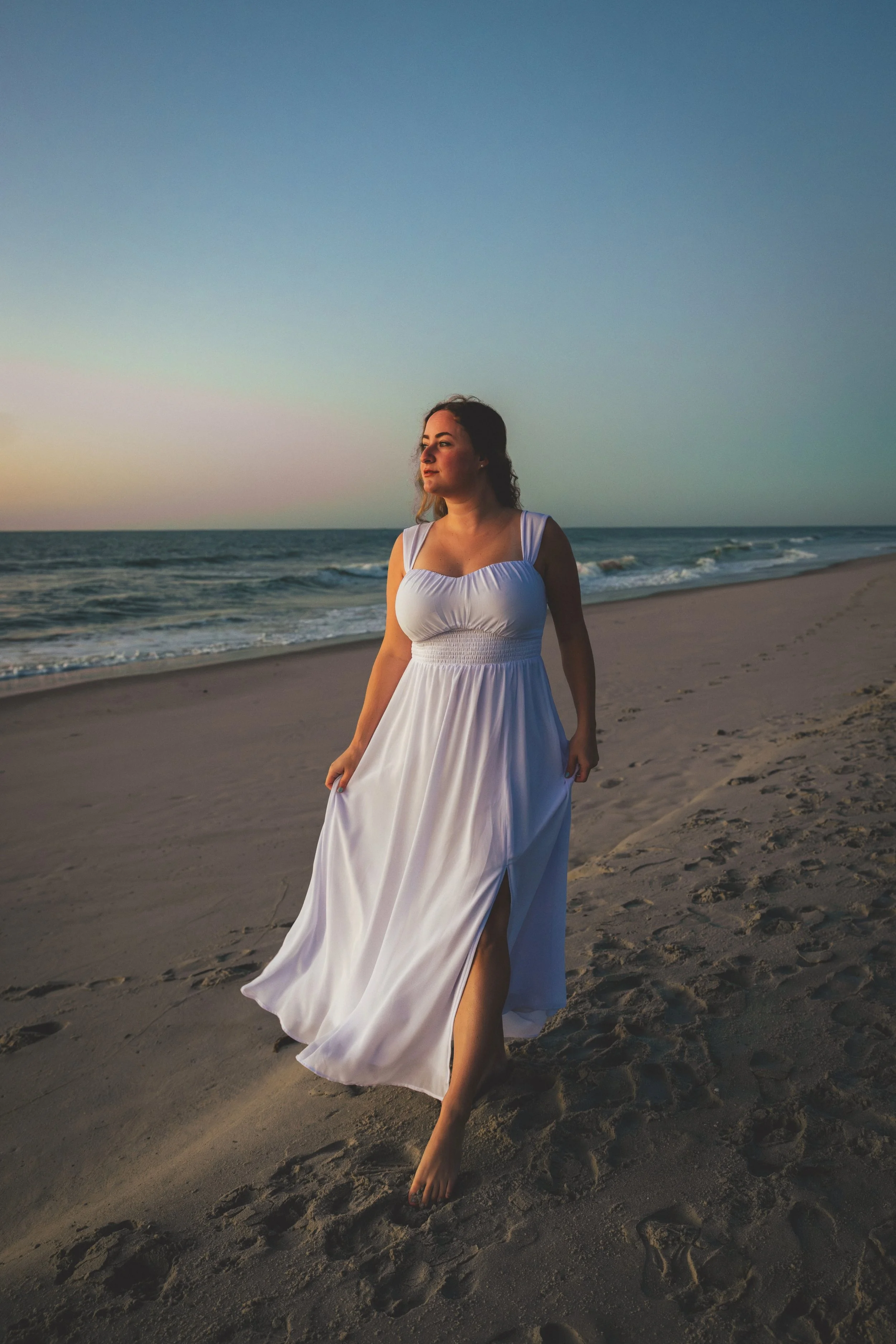 A woman in a white dress walking barefoot on the sandy beach at sunset, with the ocean and sky in the background.