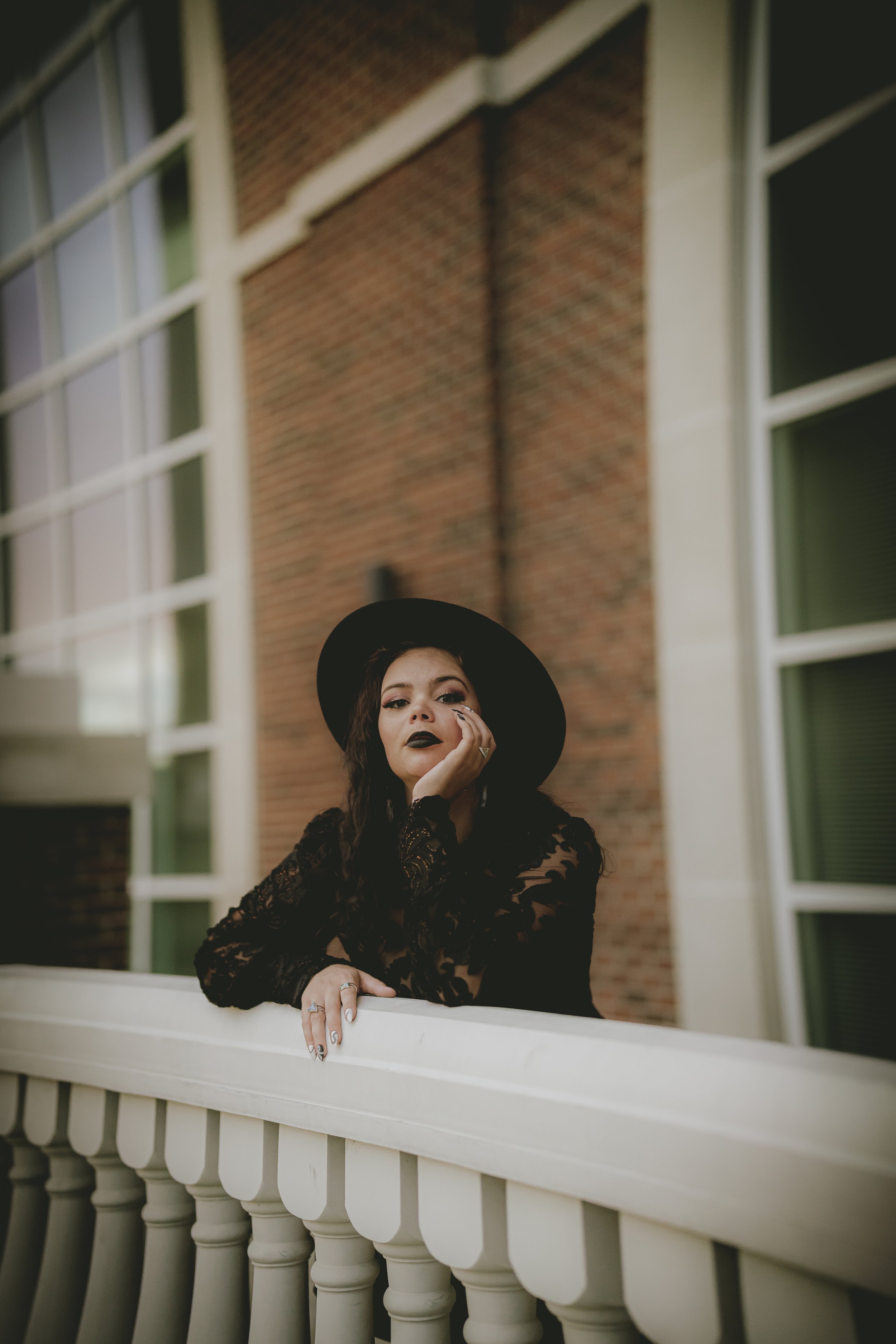 A woman with dark makeup, wearing a black lace dress and a wide-brimmed black hat, leaning on a white railing in front of a brick building.