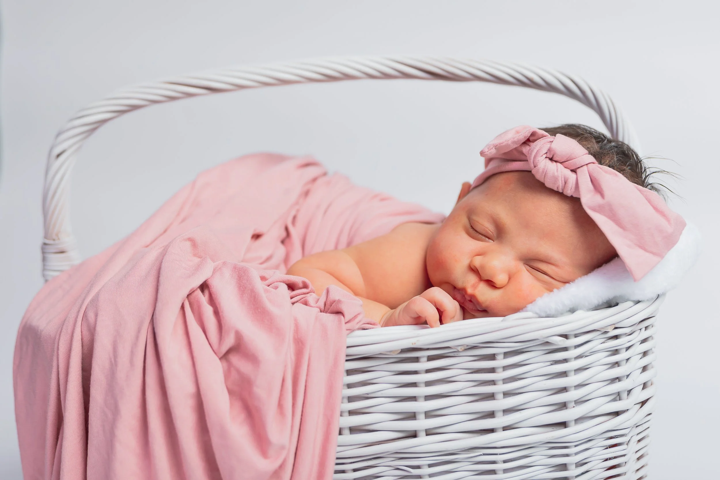 A sleeping newborn baby with a pink headband and pink blanket in a white wicker basket.