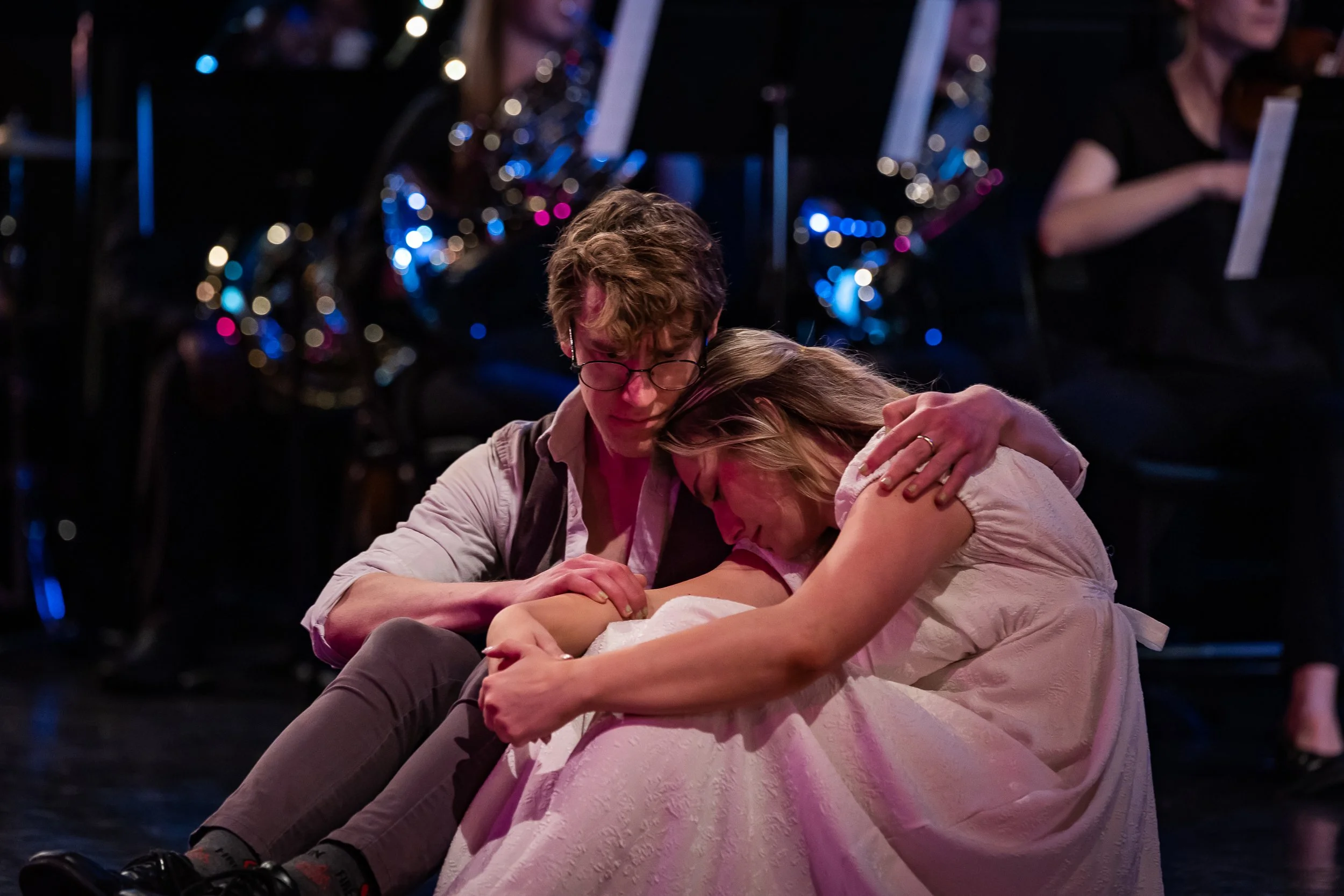 Two actors on stage embrace during a theatrical performance, with musicians in the background.