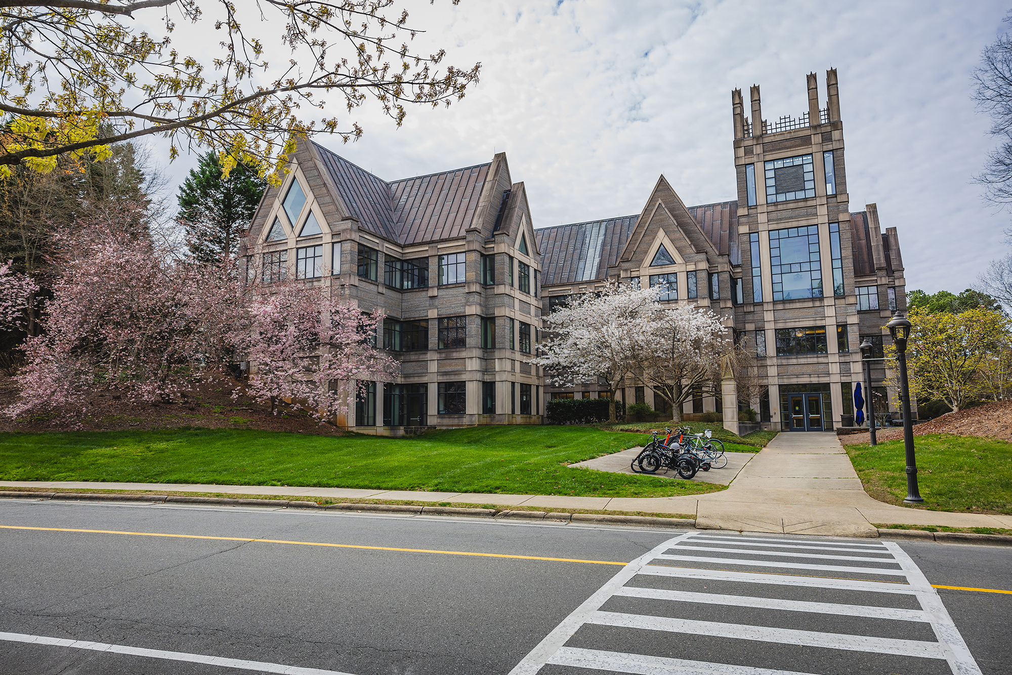 A large stone building with gothic architectural features, including pointed arches and a tower, surrounded by blooming cherry blossom trees and green grass, with a sidewalk, bicycles, and lampposts in the foreground.