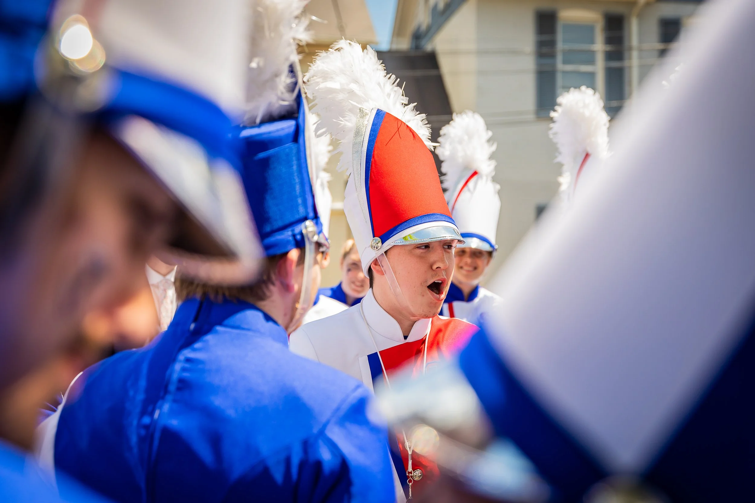 Young person in a marching band uniform with a tall red, white, and blue hat, surrounded by fellow band members in similar uniforms, outdoors in sunlight.