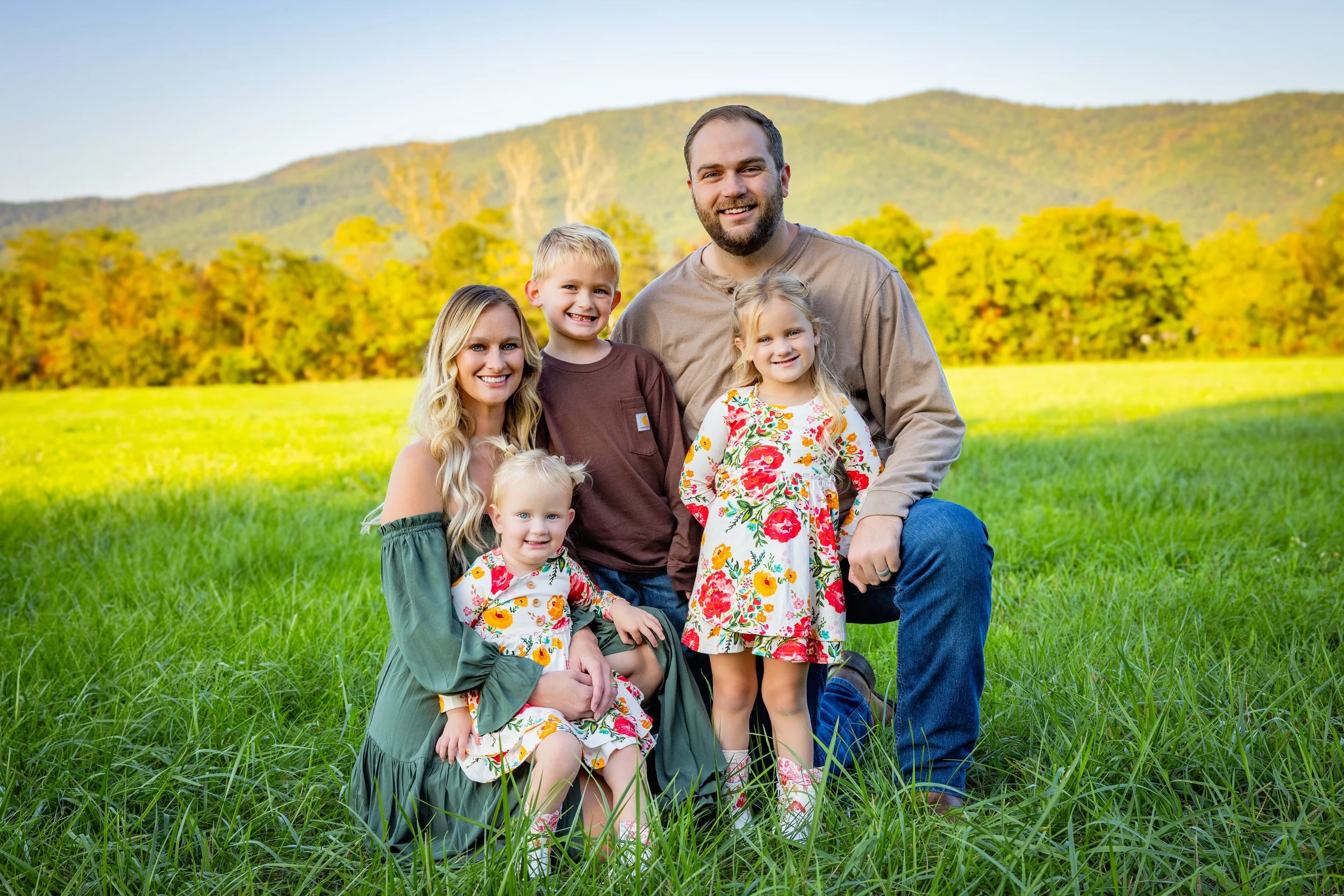 A family of six posing together outdoors in a green field with trees and mountains in the background, all smiling.