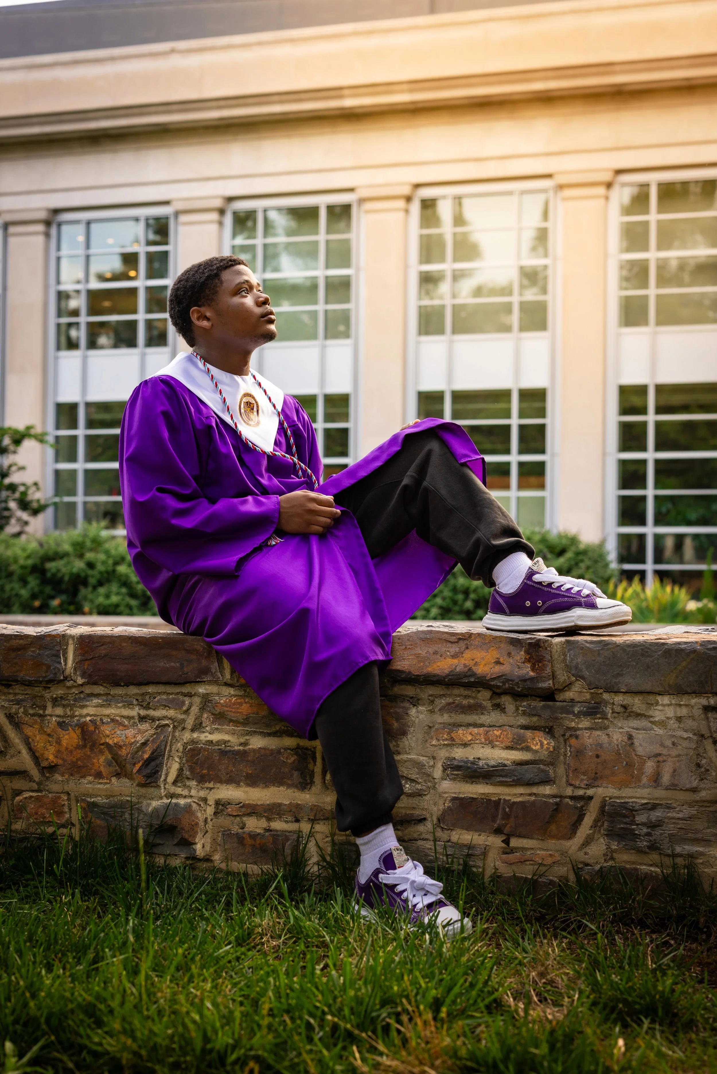 Young person in a purple graduation gown sitting on a stone wall in front of a modern building with large windows, looking upward.