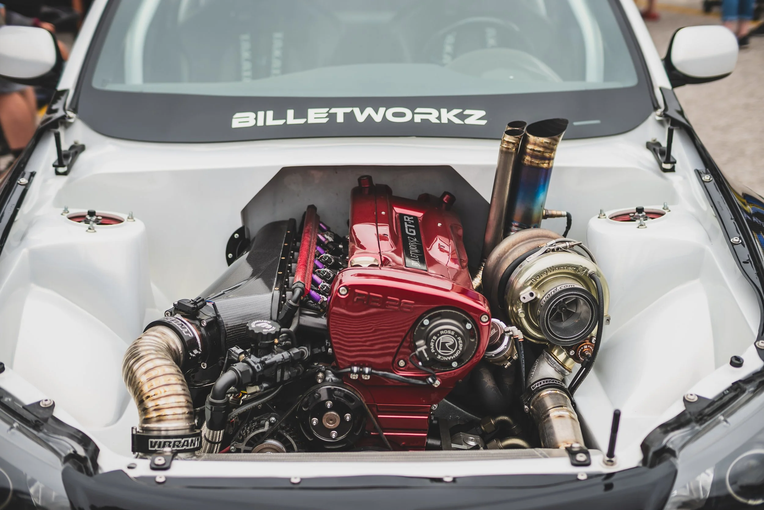 Car engine with a red and black turbocharger, visible piping, and components, in a car with a white interior and a sticker that reads 'BILLETWORKZ' on the dashboard.