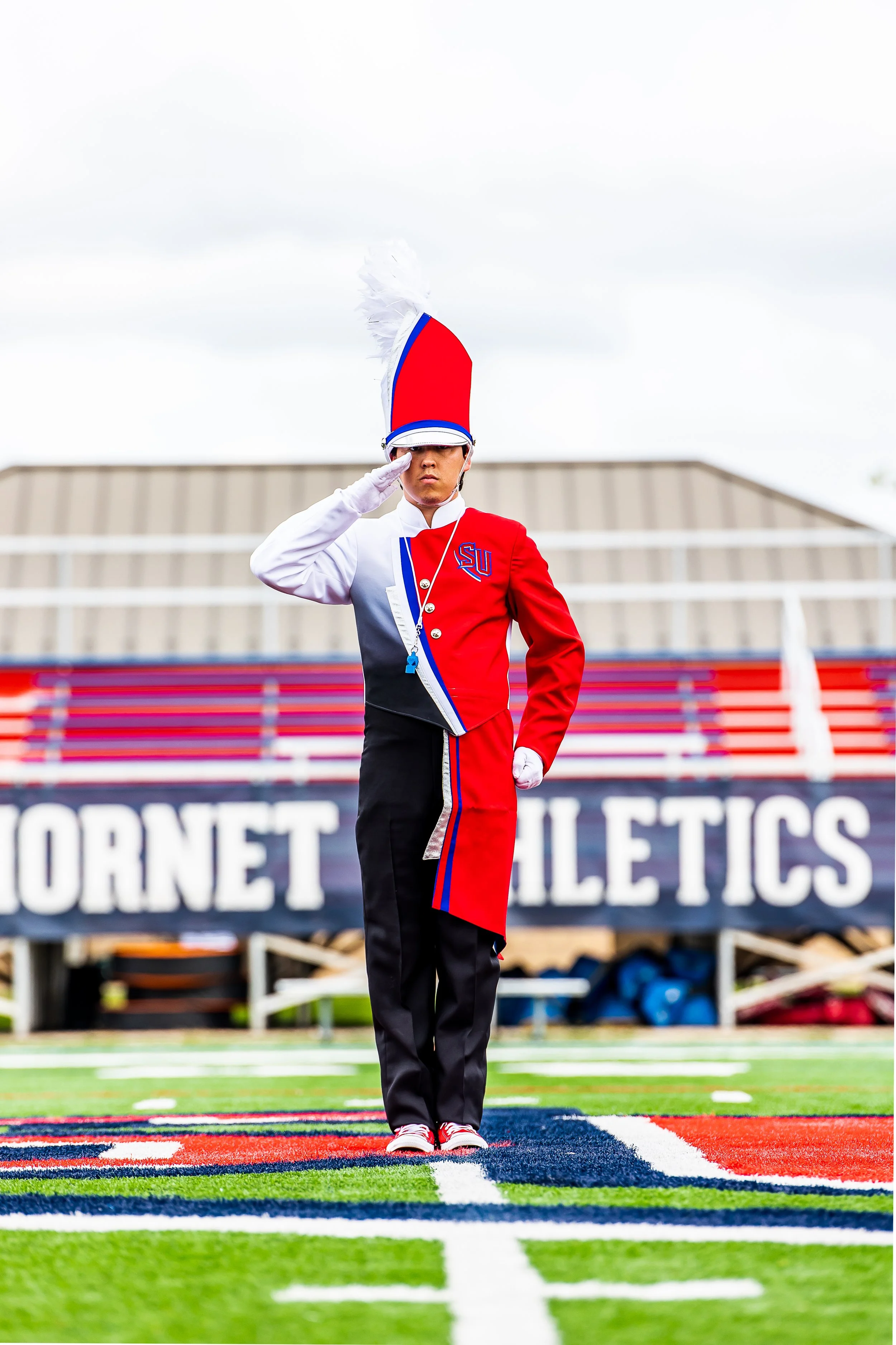 A marching band member in a red, black, and white uniform, wearing a tall feathered hat, standing at attention and saluting on a football field with a Hornet Athletics banner in the background.