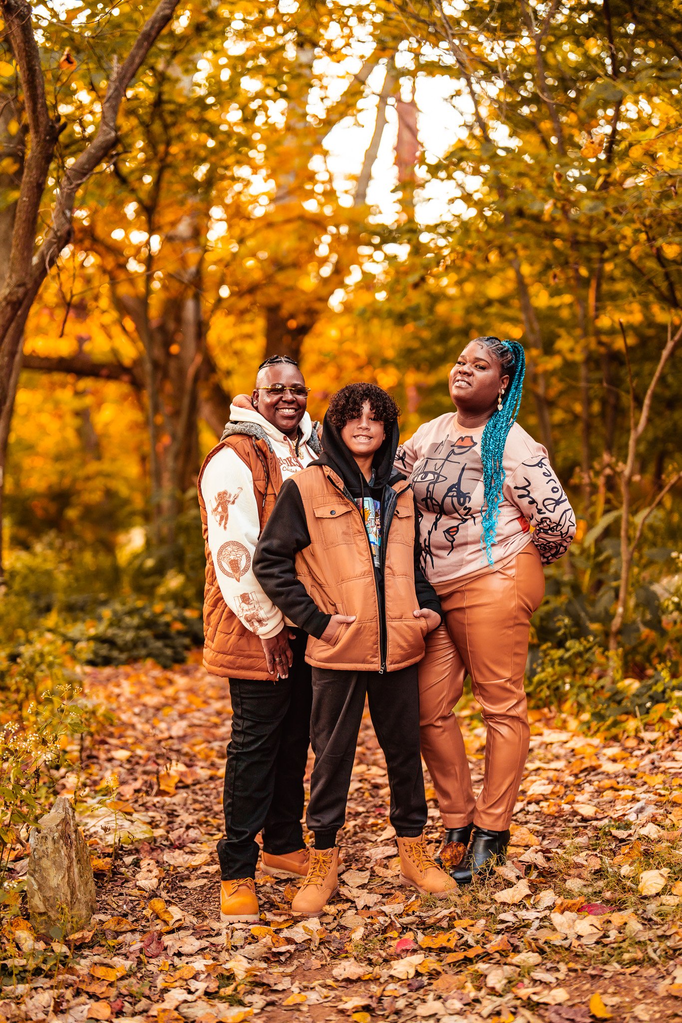 Three people standing on a leaf-covered trail in a wooded area with autumn foliage. They are smiling and wearing fall clothing, including jackets and boots.