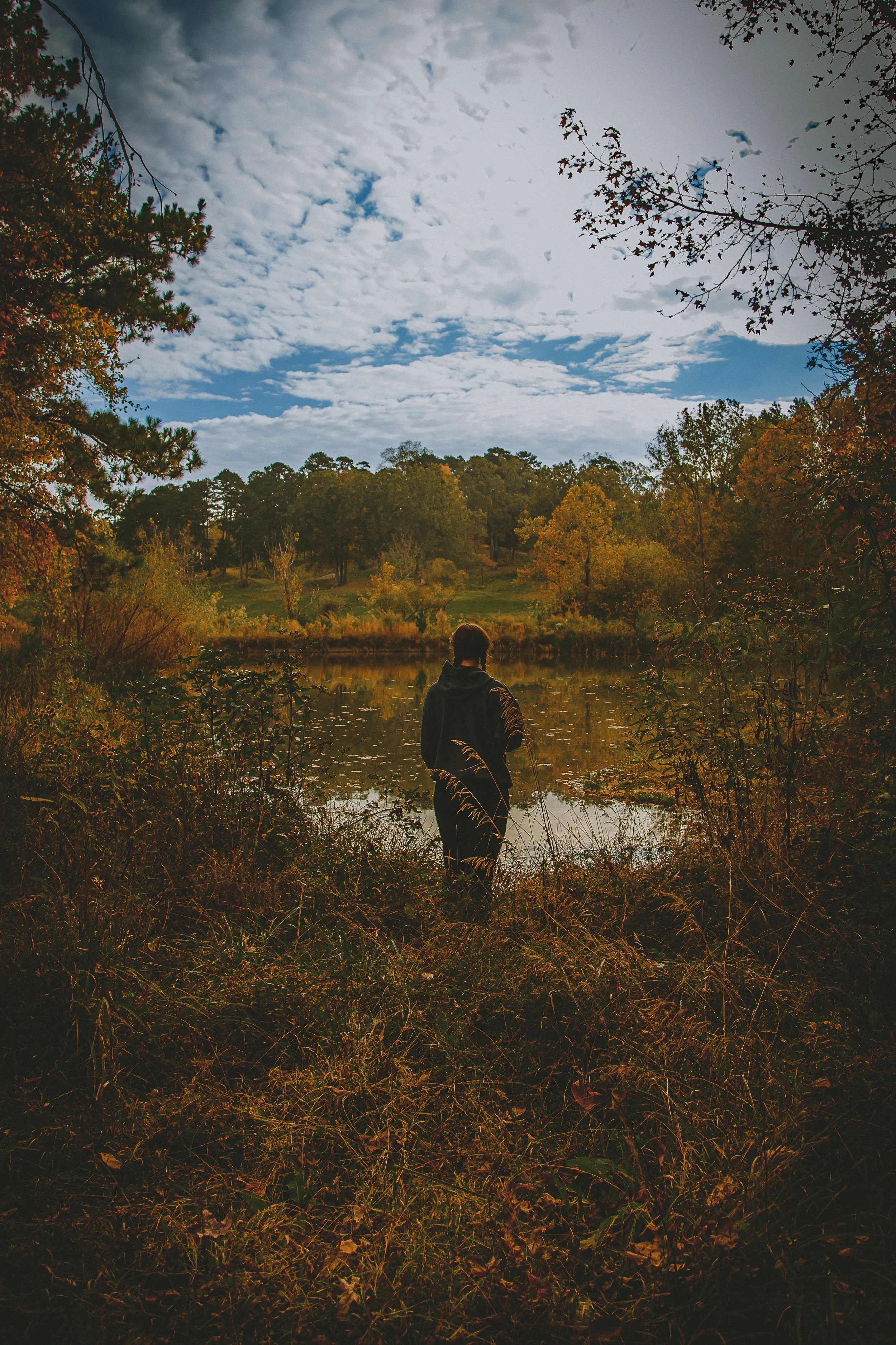 Person standing by a lake during fall, surrounded by trees with autumn foliage, under a partly cloudy sky.