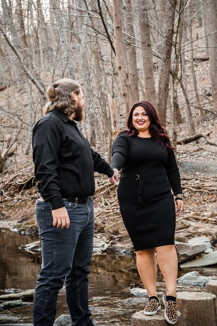 A man and a woman holding hands and smiling in a wooded outdoor setting near a small creek, with trees and rocks in the background.