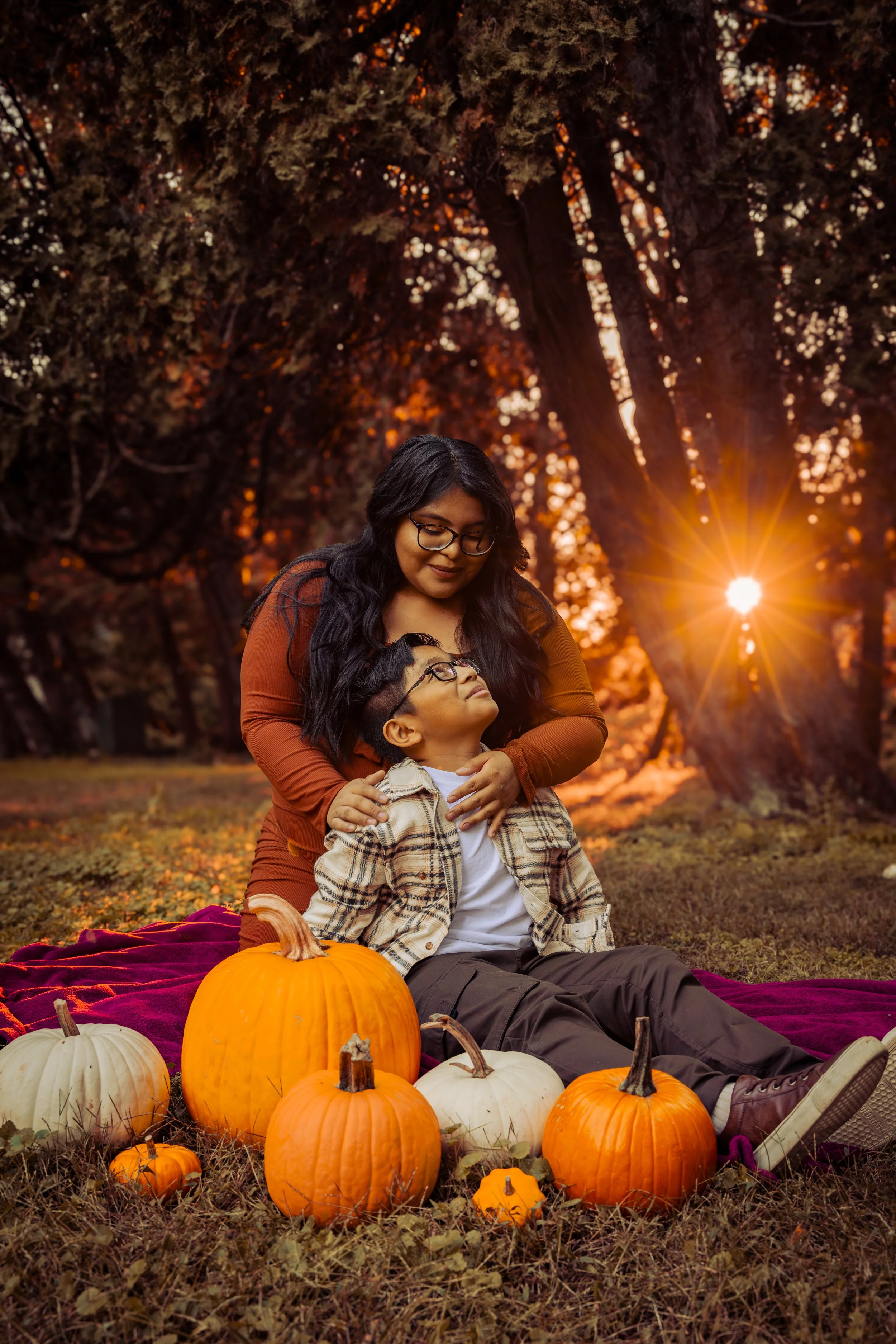 A woman and a boy sitting on the ground surrounded by pumpkins in an outdoor setting during sunset