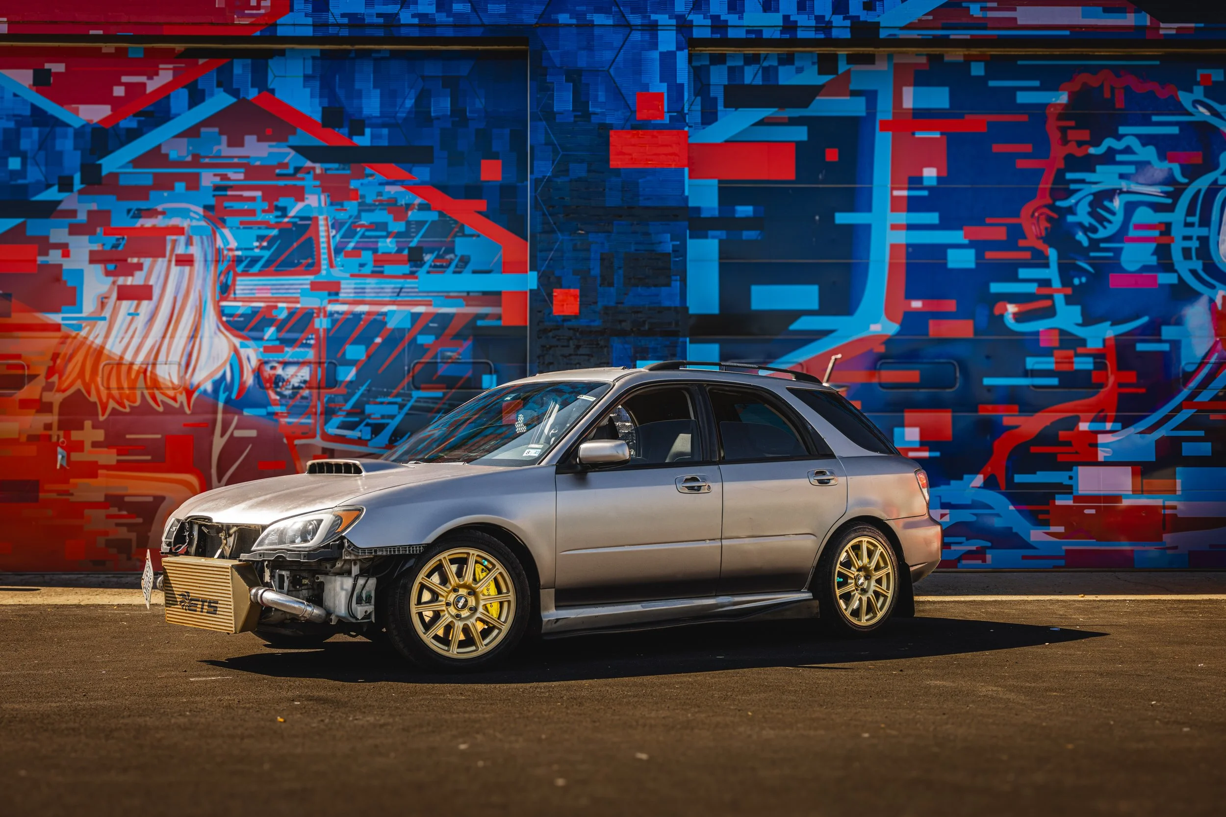 A silver car with a damaged front end and gold wheels parked in front of a vibrant, abstract street art mural with blue and red geometric designs and stylized figures.