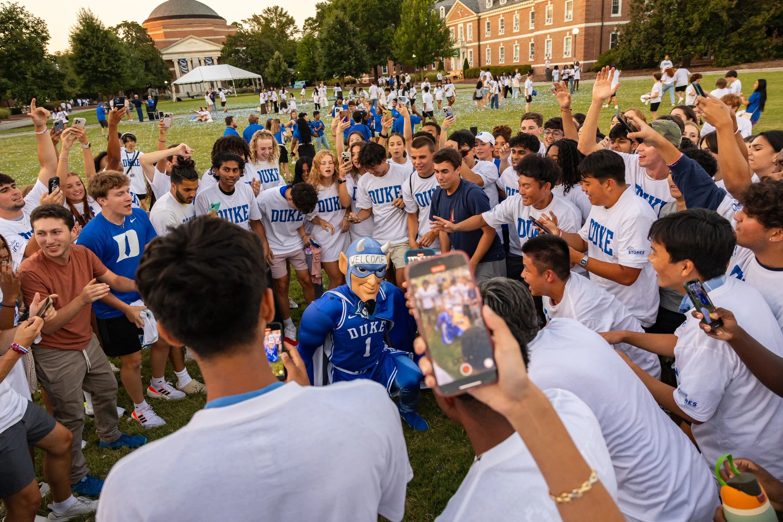 Group of students gathered outdoors on a university campus, taking photos and videos of a mascot dressed in Duke University gear, surrounded by friends and classmates.