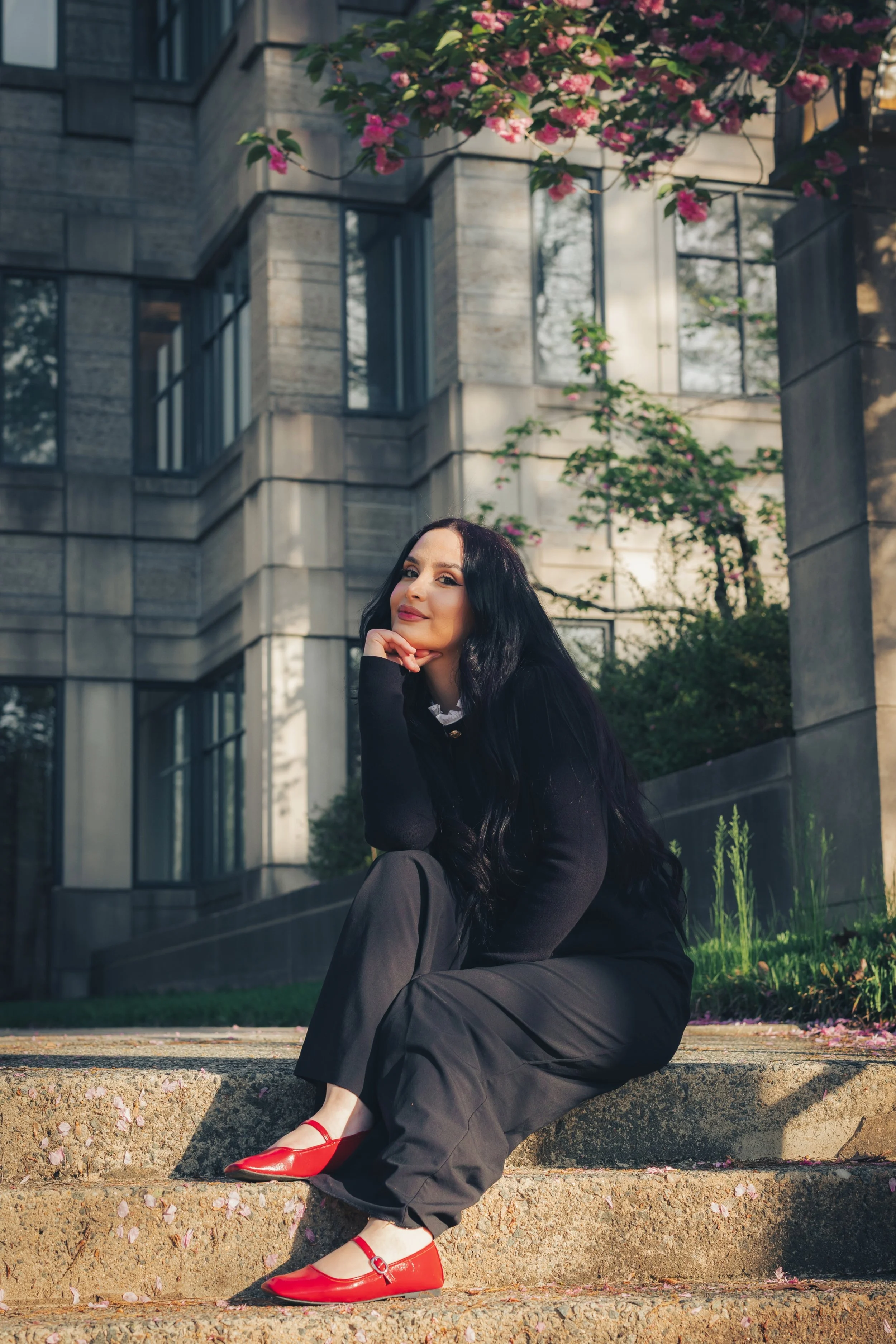 A woman with long black hair, wearing a black sweater, black pants, and red shoes, sitting on outdoor concrete steps near a building covered with pink flowering bushes.