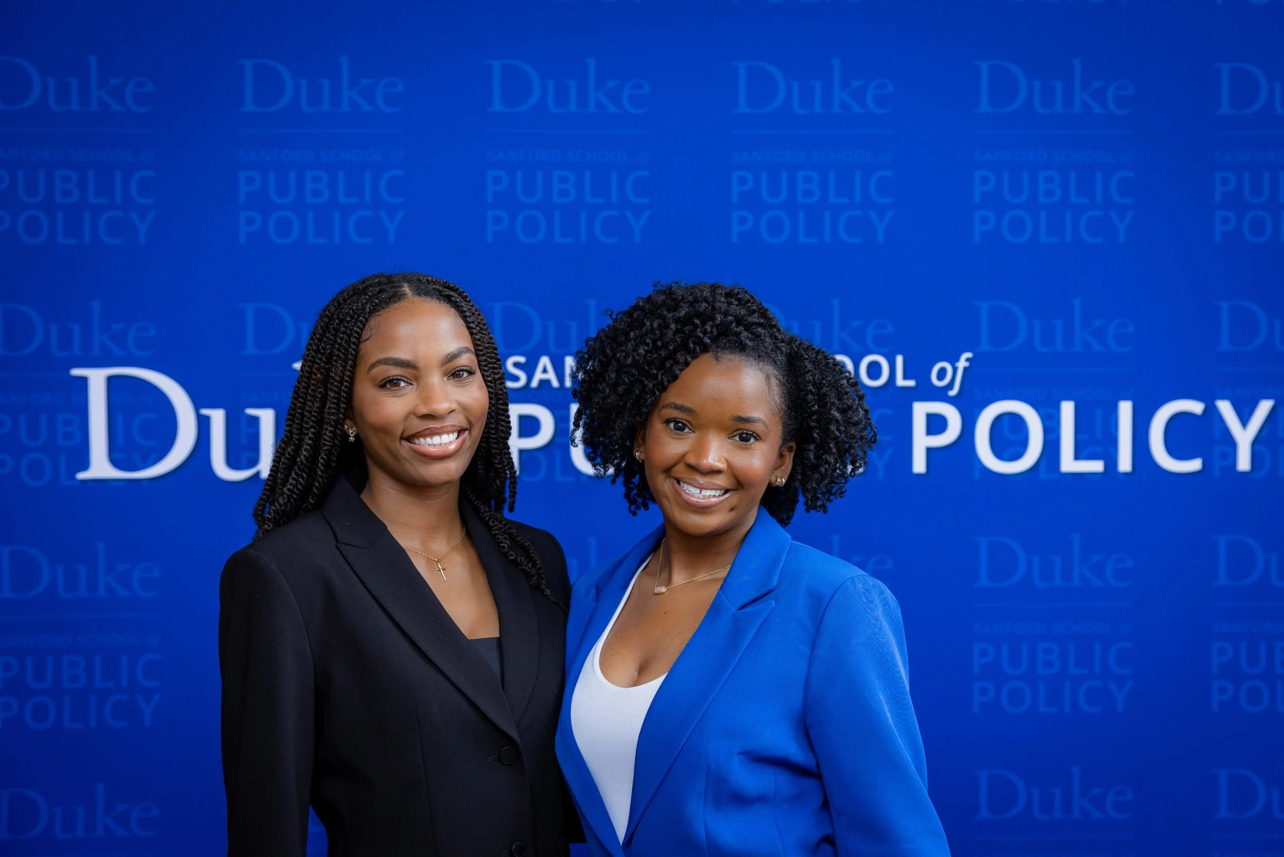 Two women standing in front of a blue backdrop with 'Duke' and 'Public Policy' text, smiling at the camera.