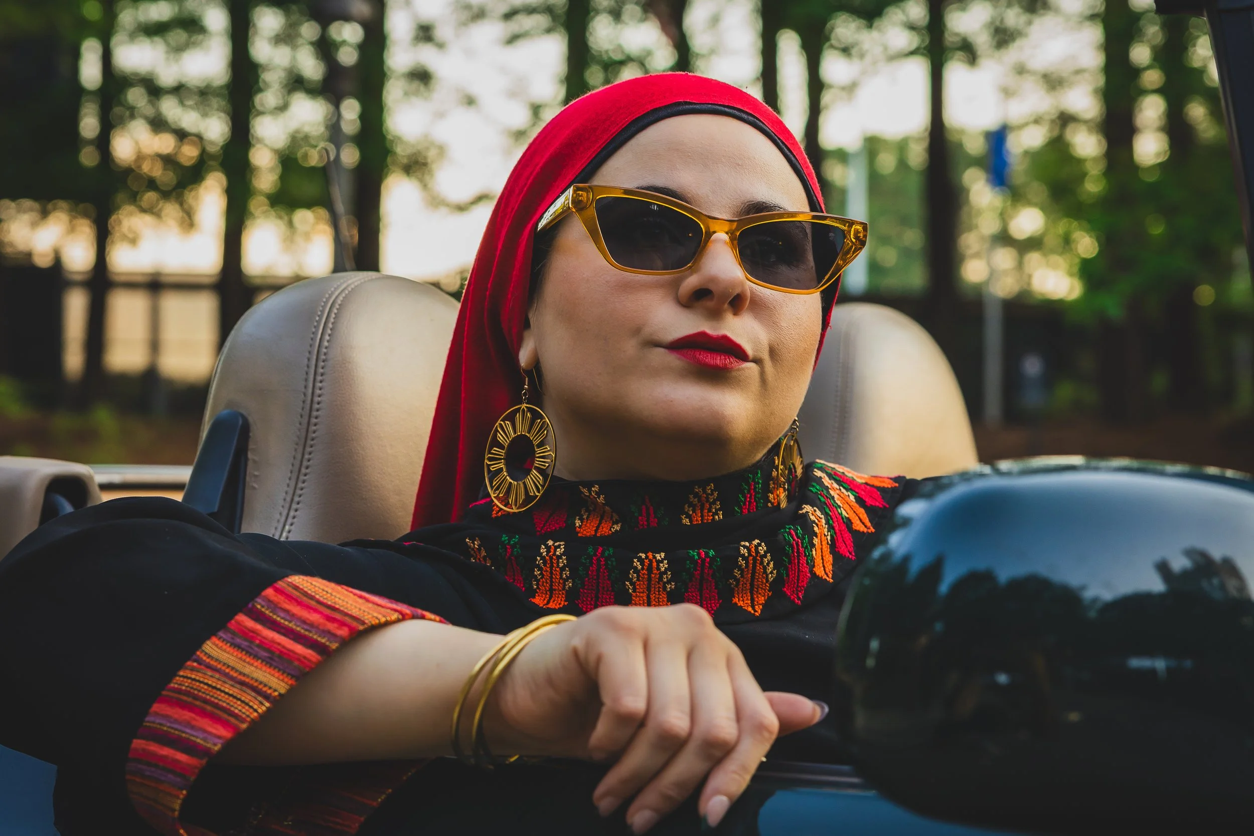 A woman with red lipstick, wearing sunglasses, earrings, a black embroidered top, and a red headscarf, sitting in a vintage car with a forest background.