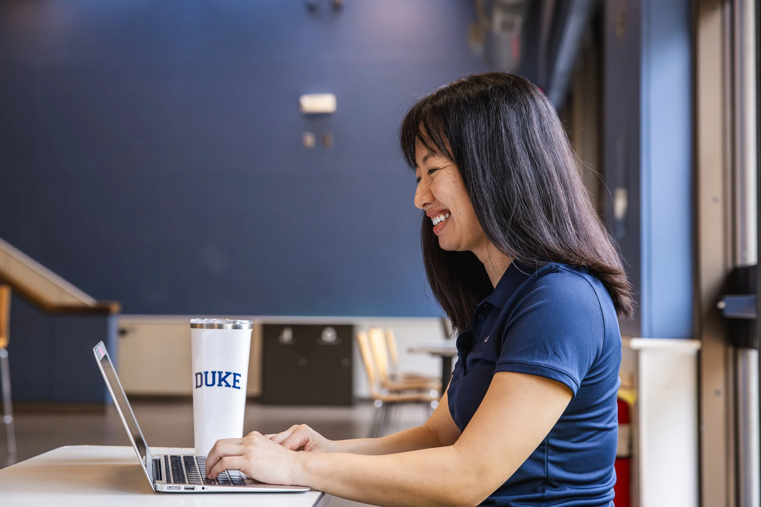 A smiling woman with dark hair working on a laptop at a table with a Duke University tumbler next to her in a bright room.