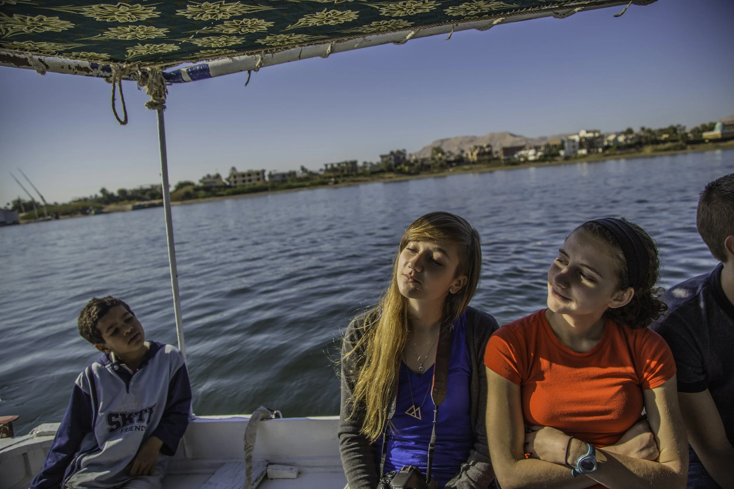Four young people sitting on a boat on a lake, with a city and mountains in the background. The sun is shining, and the group appears to be relaxing or enjoying the ride.