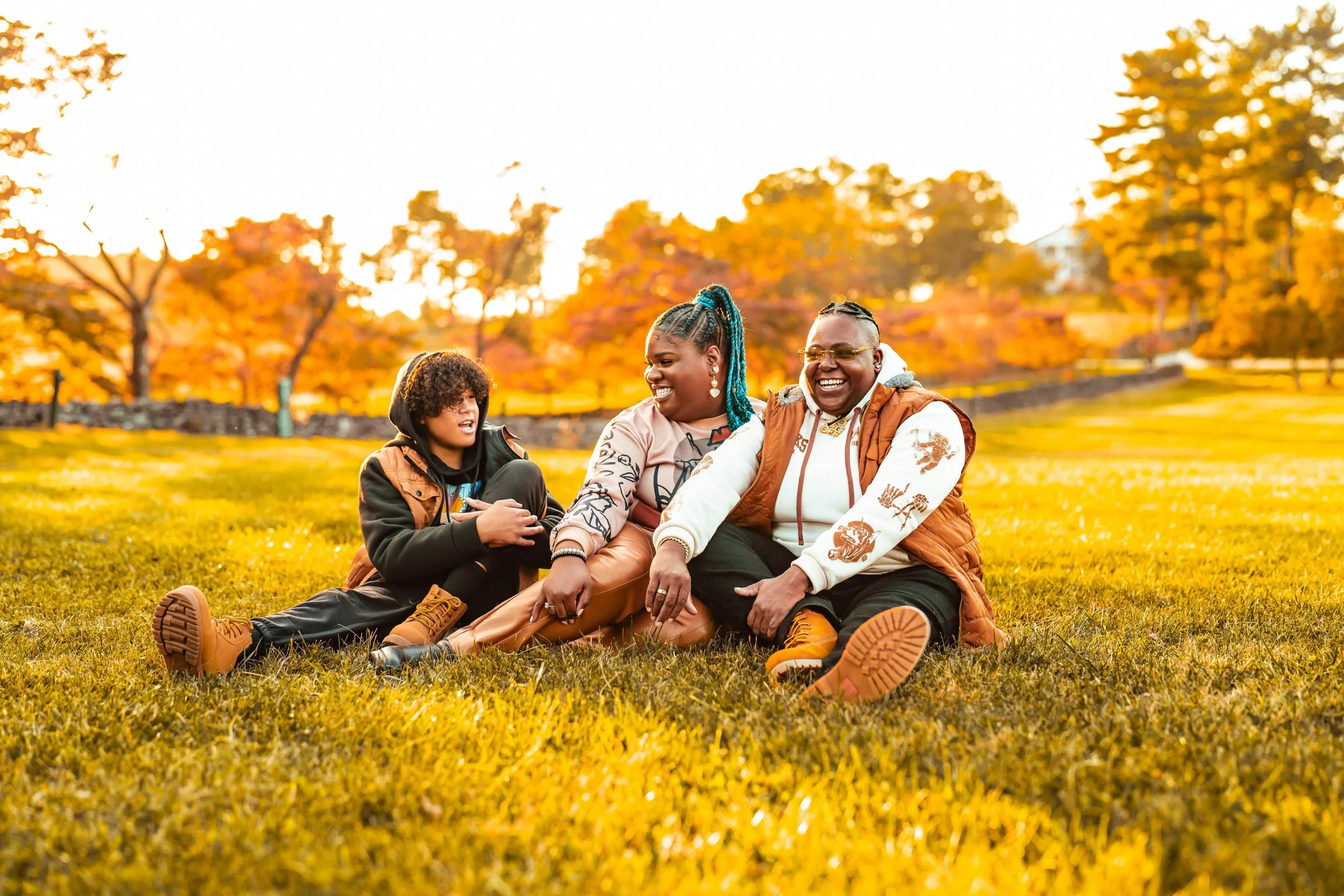 Three friends sitting on the grass in a park during autumn, smiling and enjoying the warm sunlight with colorful fall trees in the background.