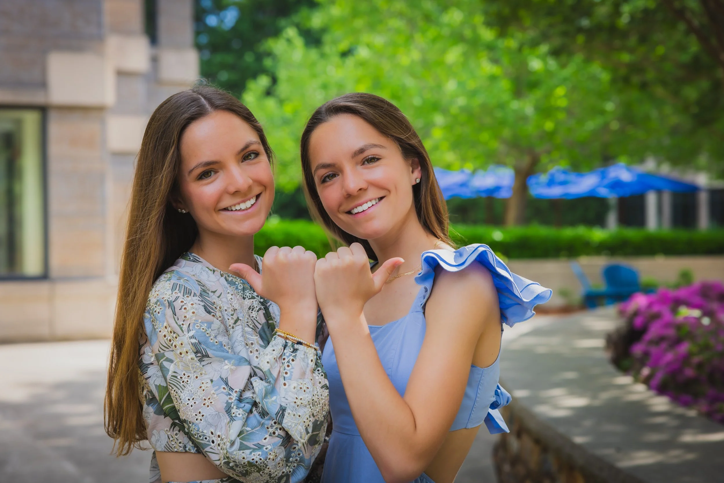 Two young women with long brown hair smiling and making a friendly gesture with their arms around each other outdoors with greenery and purple flowers in the background