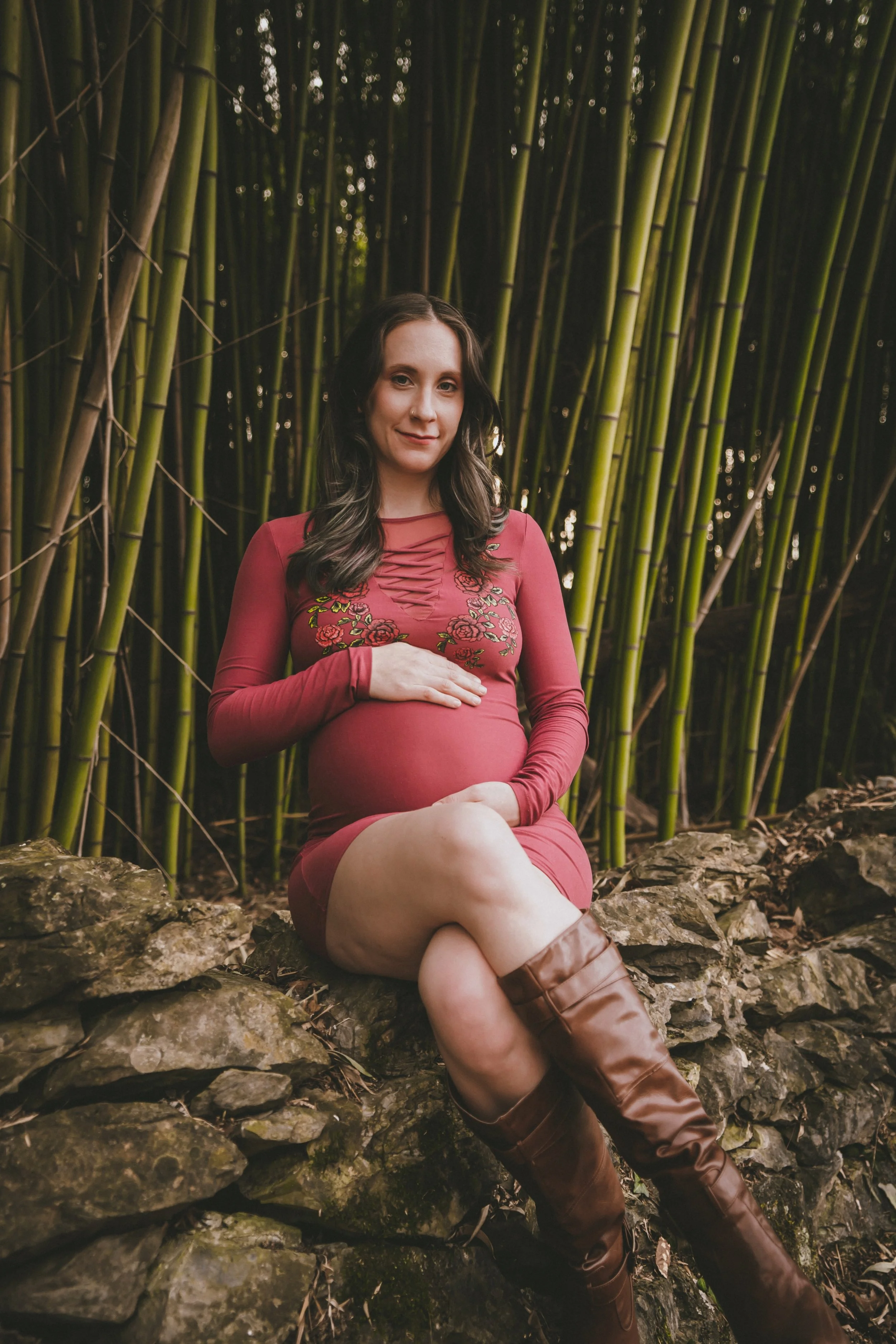A pregnant woman sitting on a rock wall in front of tall bamboo stalks, wearing a red dress and brown knee-high boots, looking at the camera and touching her baby bump.