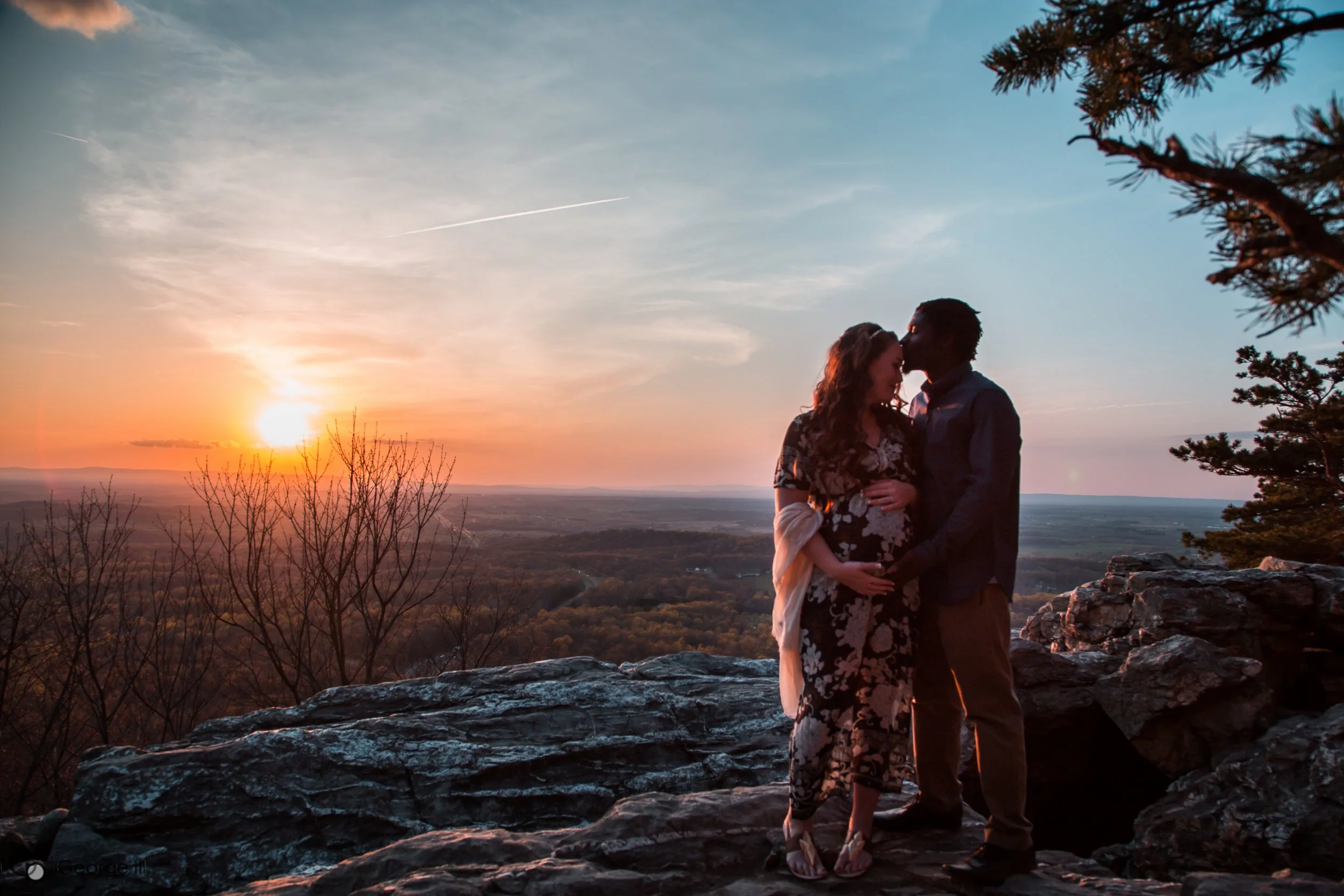 A couple stands on rocks at sunset, with the woman touching her pregnant belly as they share a kiss. The sunset and a scenic landscape of trees and distant hills are visible in the background.
