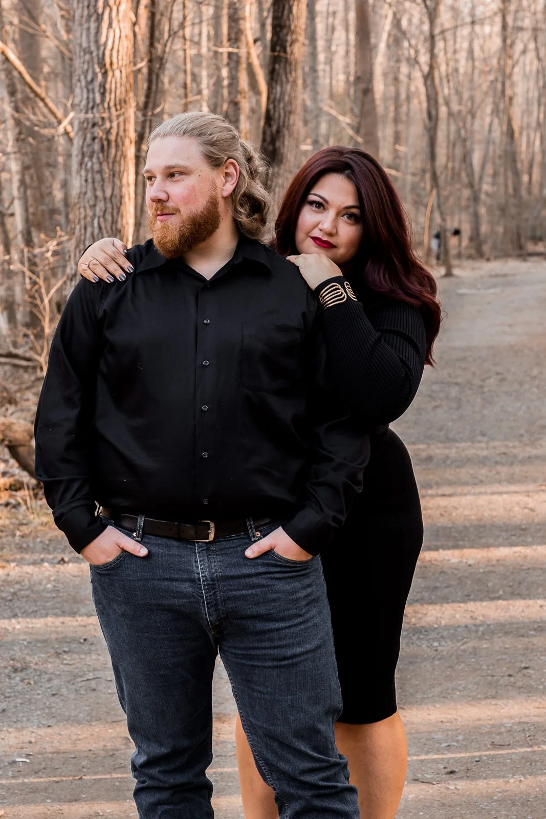 A man and woman are standing outdoors in a wooded area during autumn. The man is wearing a black button-up shirt and jeans, with his hands in his pockets, and looks to the side. The woman is wearing a black dress and has her arms resting on the man's