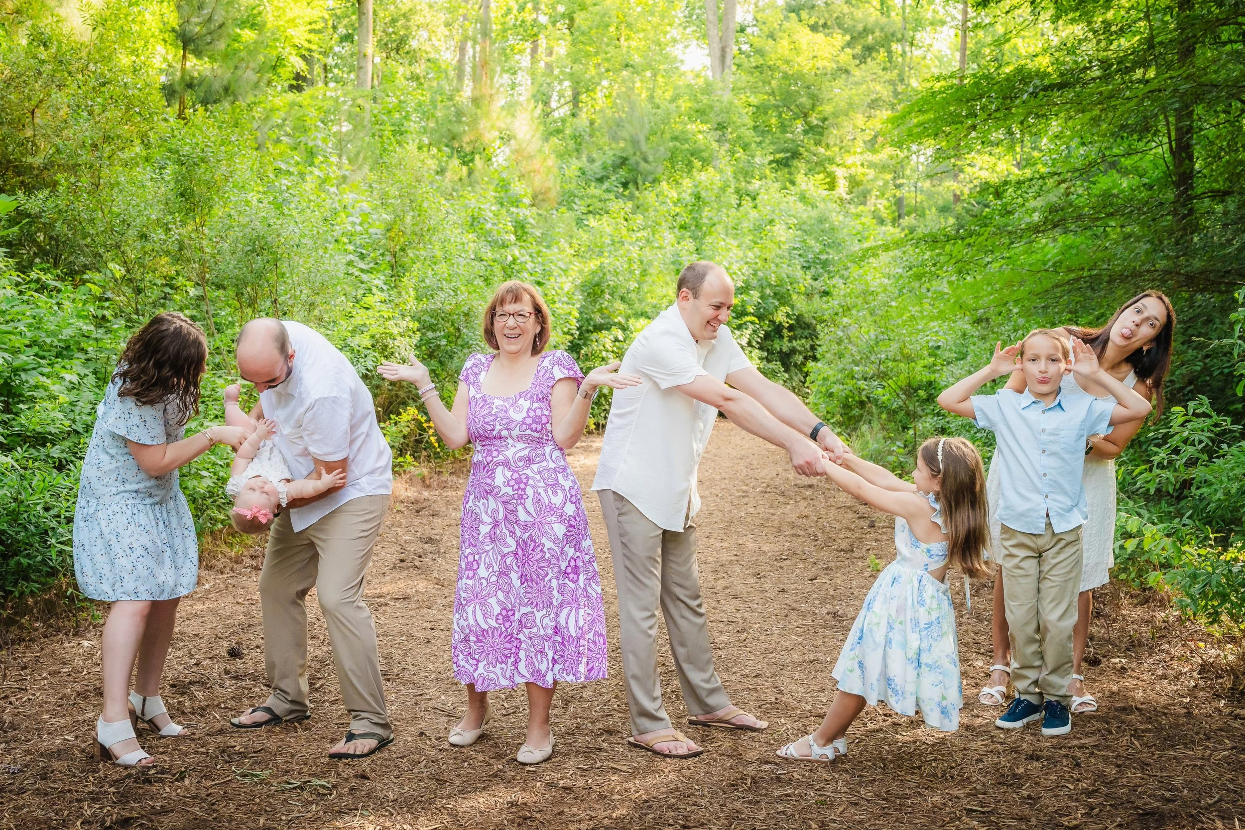 A family of eight people, including children and adults, holding hands and making funny faces while standing and playing in a lush, green forest.
