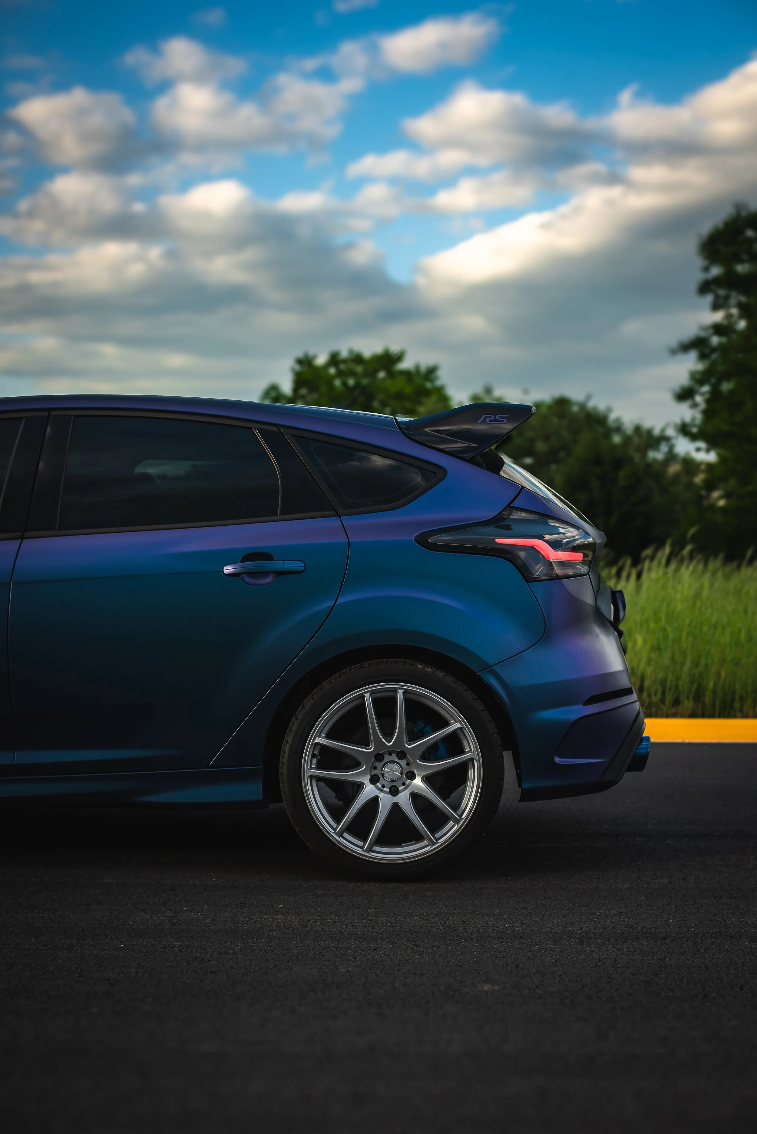 Close-up of the rear side of a blue sports car parked on a road with trees and a partly cloudy sky in the background.