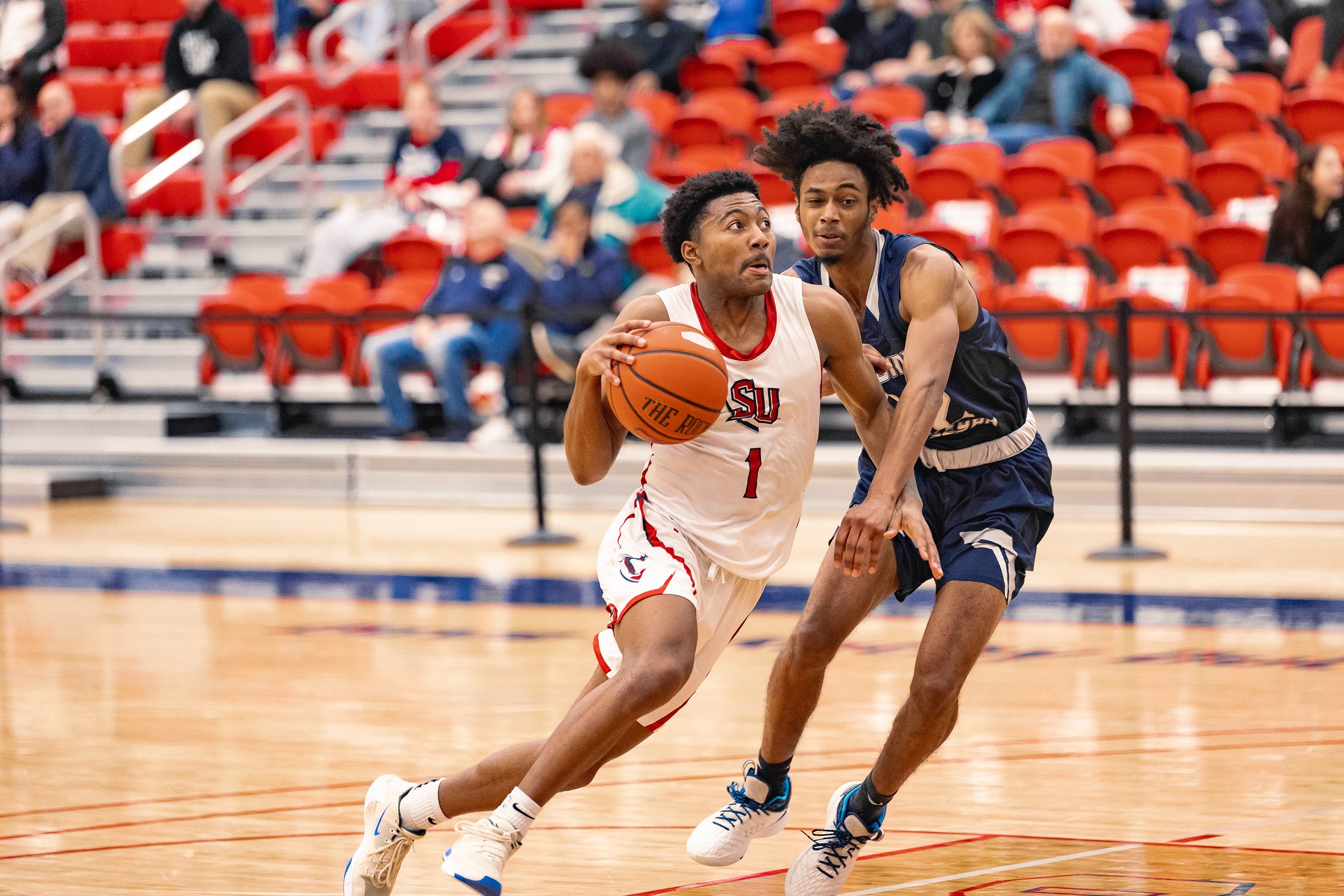 Two young men playing basketball on an indoor court, one in a white jersey with the number 1 holding the ball, the other in a dark navy jersey defending.