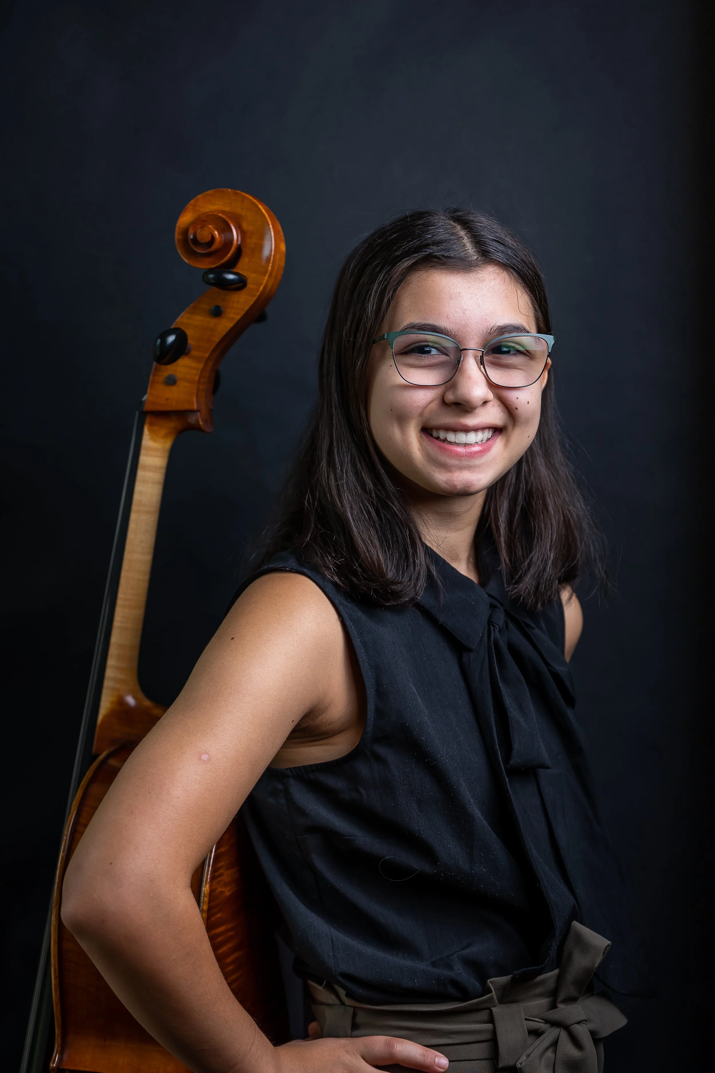 A young woman with long dark hair and glasses smiling while posing with a wooden cello, dressed in a sleeveless black top, against a dark background.
