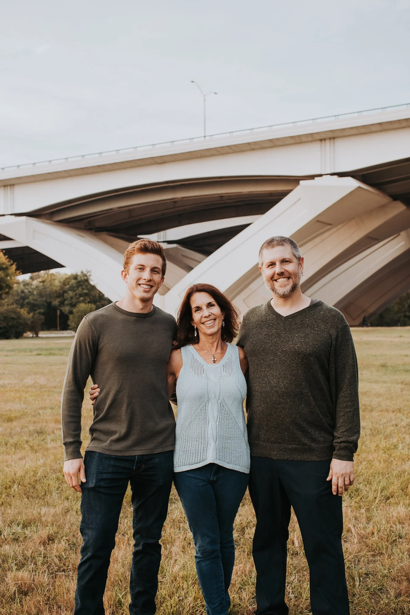 A family of three standing together outdoors under a bridge, smiling at the camera. The family consists of a young man, a middle-aged woman, and a middle-aged man.