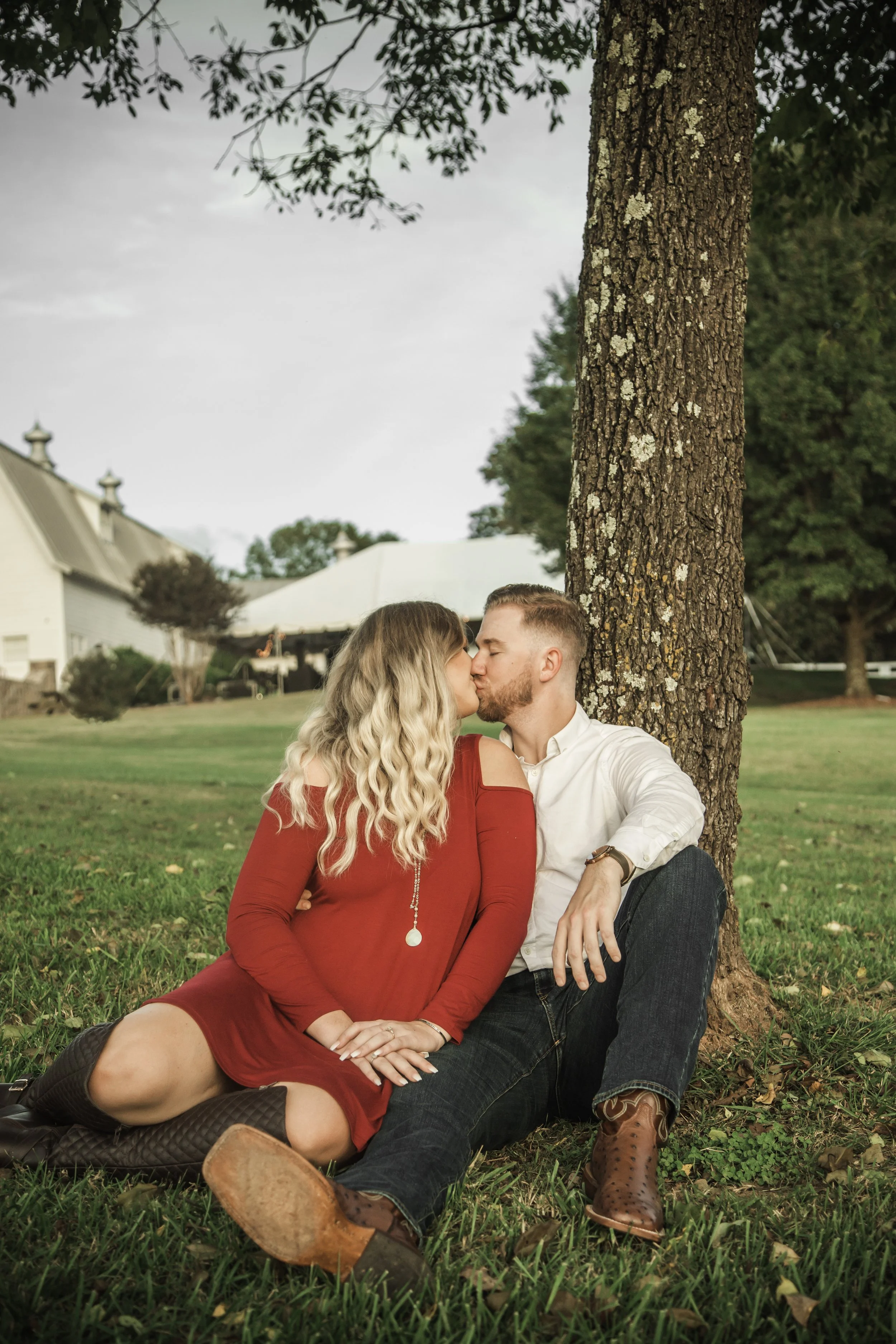 A couple kissing while sitting on grass under a large tree in a park. The woman is wearing a red dress and the man a white shirt and dark jeans.
