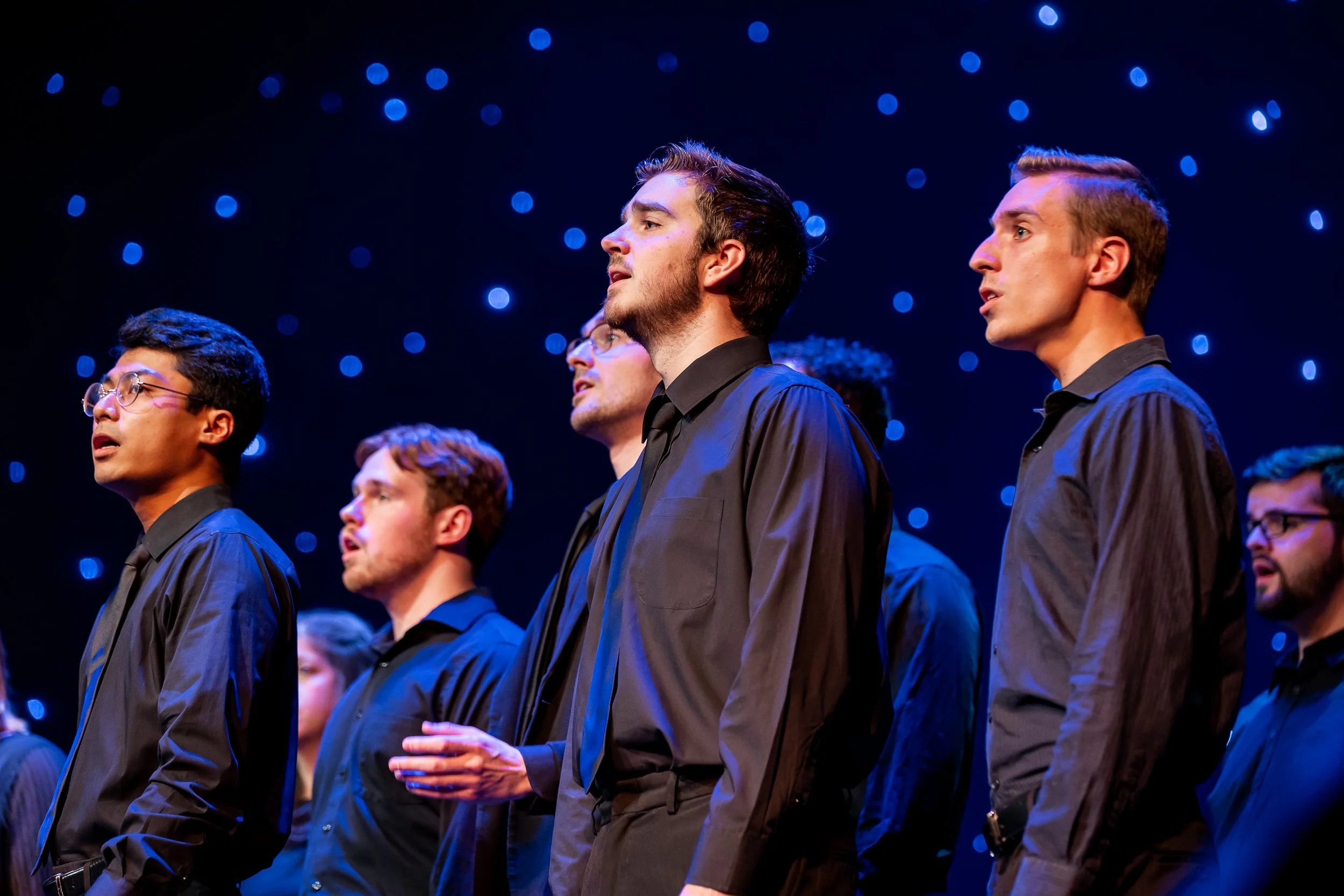 Group of male singers in black shirts singing on stage with blue starry background.
