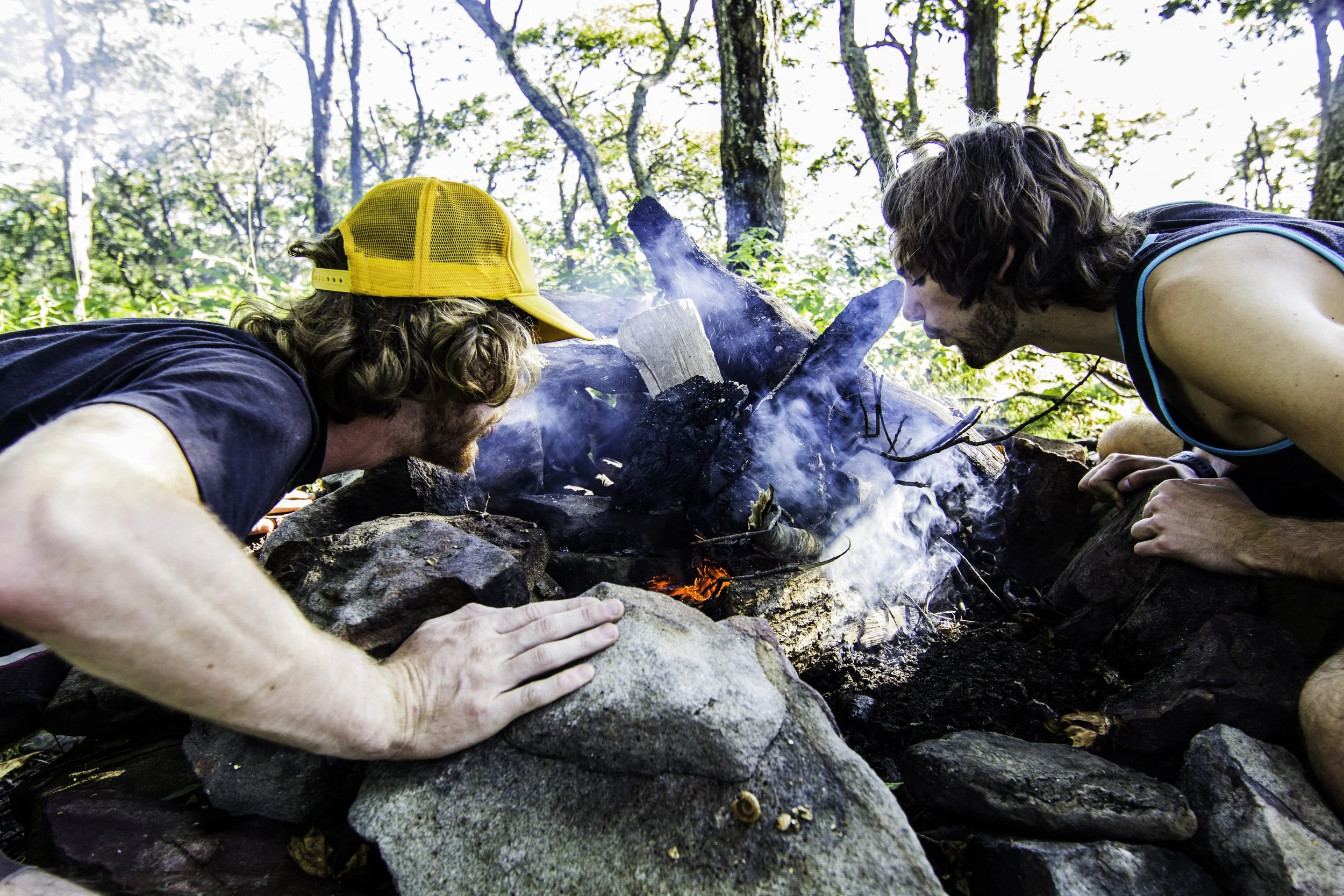 Two men lean over a campfire in a wooded area, closely observing the flames and smoke.