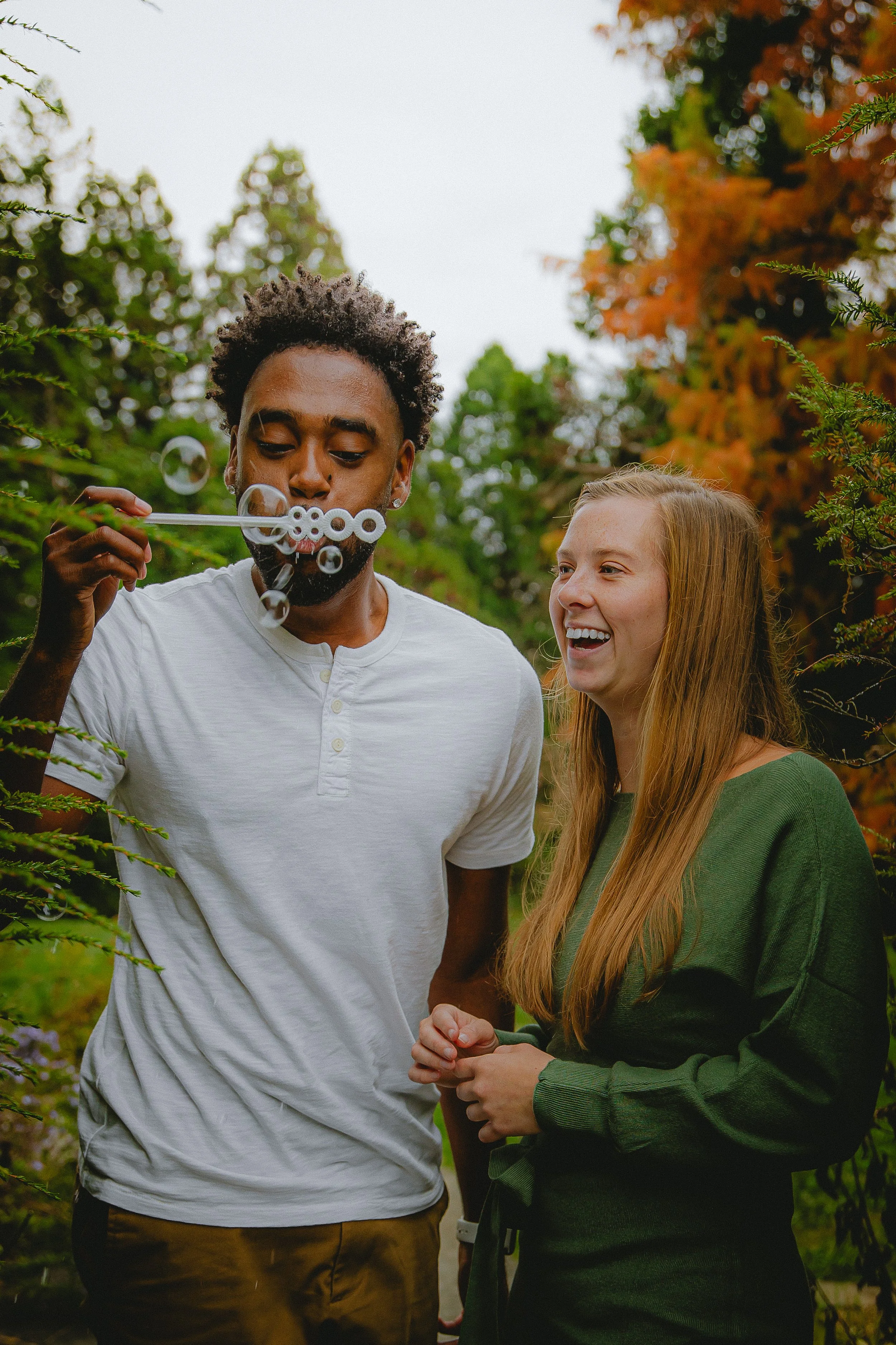 A young man blowing bubbles while a smiling young woman watches in an outdoor setting with trees and foliage.