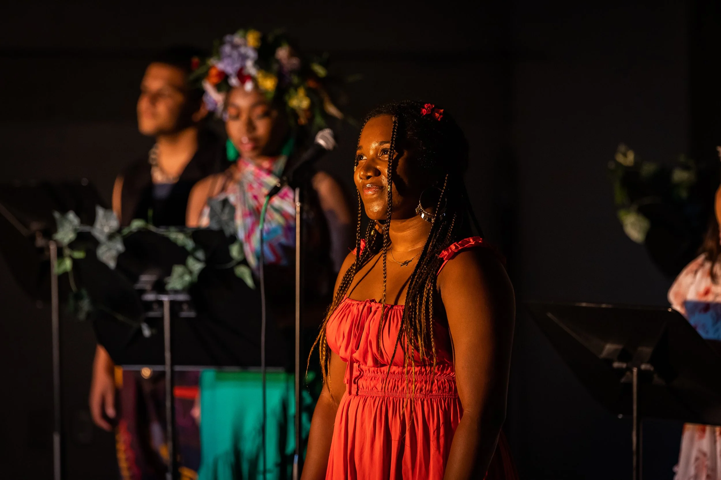 A young woman with braided hair and hoop earrings wearing a red dress standing on stage, with a woman behind her wearing a colorful flower crown and a black dress, and a man in the background in a black suit.