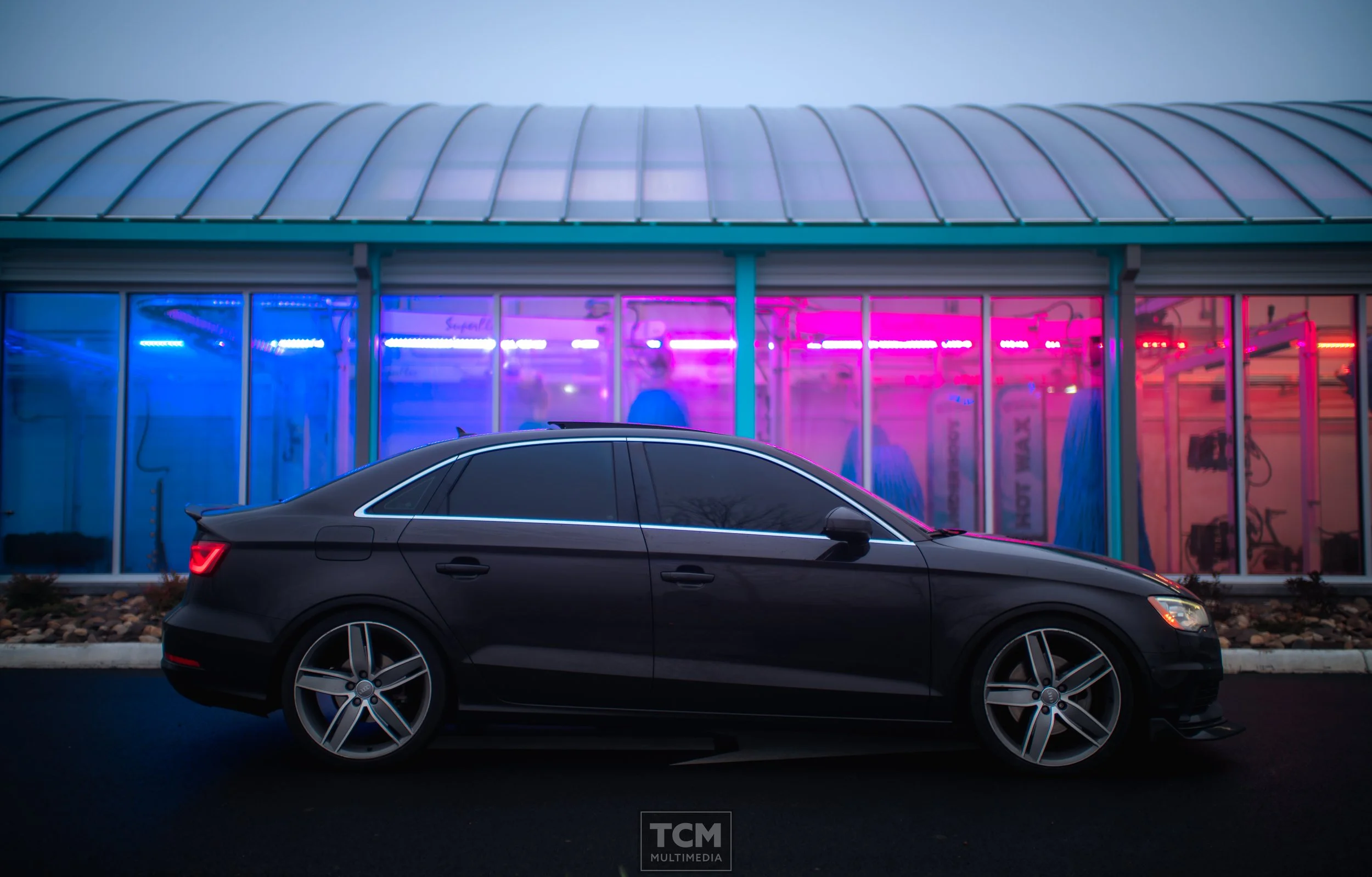 A black car parked in front of a building with glass walls illuminated by pink and purple neon lights.