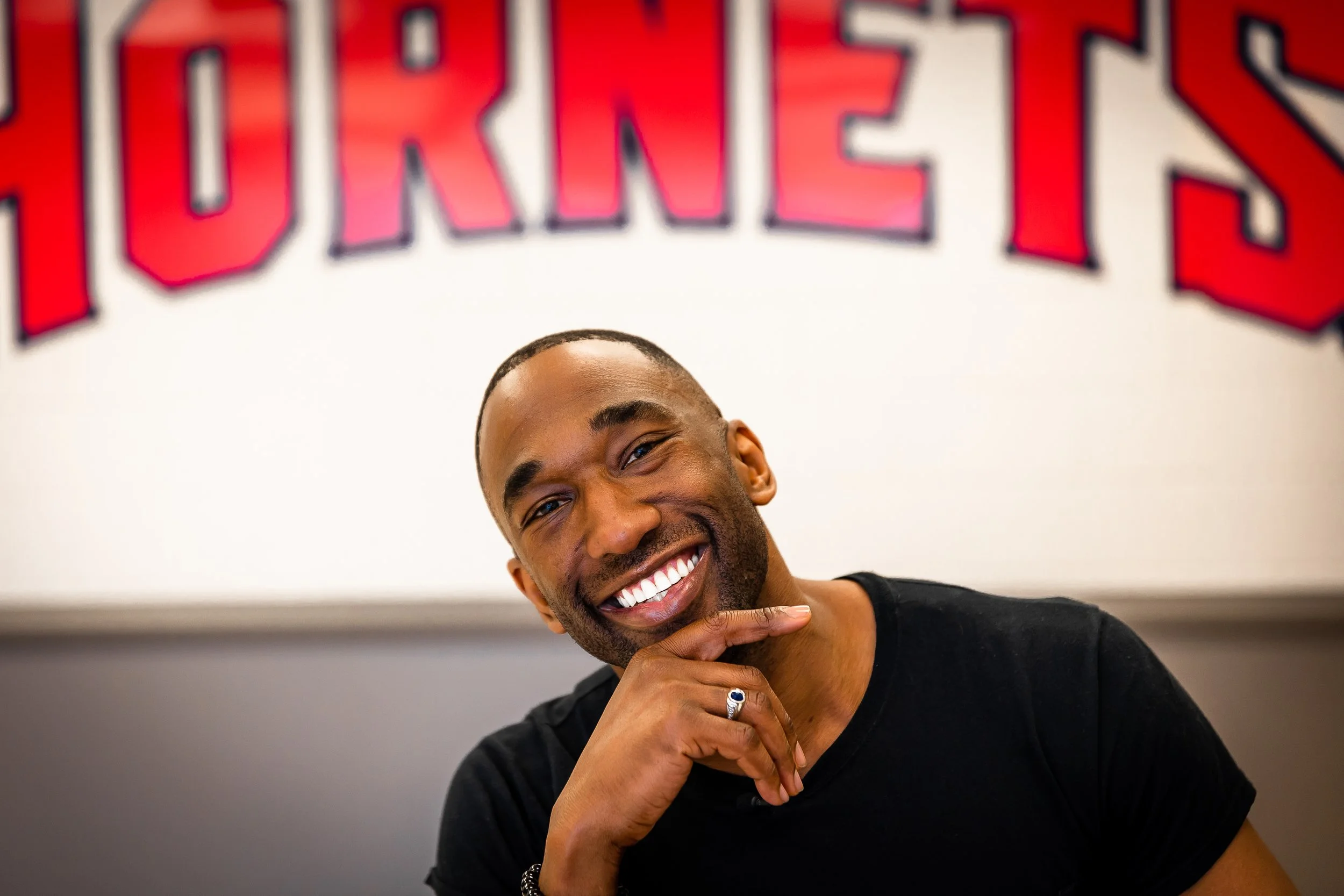 A smiling man with a beard and short hair, wearing a black shirt, resting his chin on his hand, with a large red and black 'TURTLES' sign in the background.