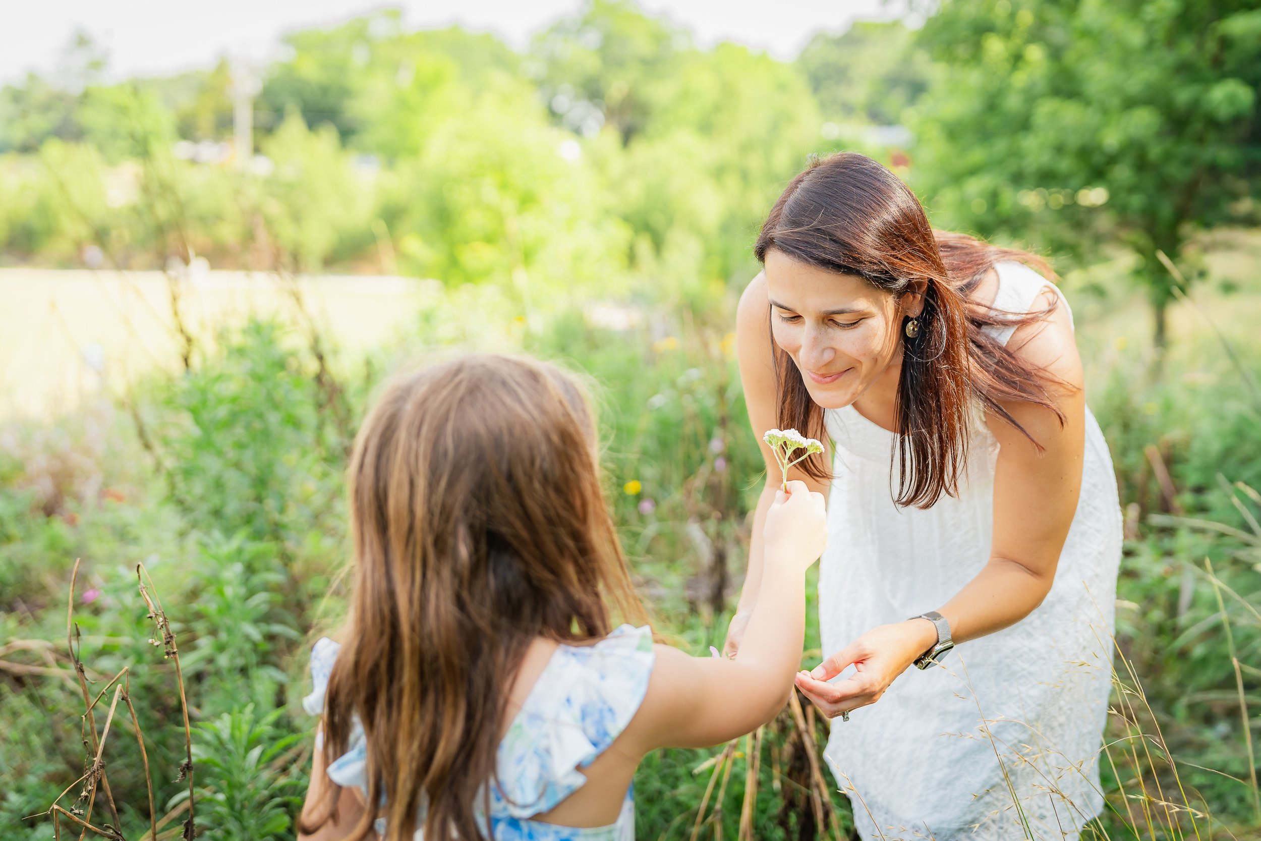 A woman with long brown hair smiling and holding a small flower, engaging with a young girl with brown hair in a garden.