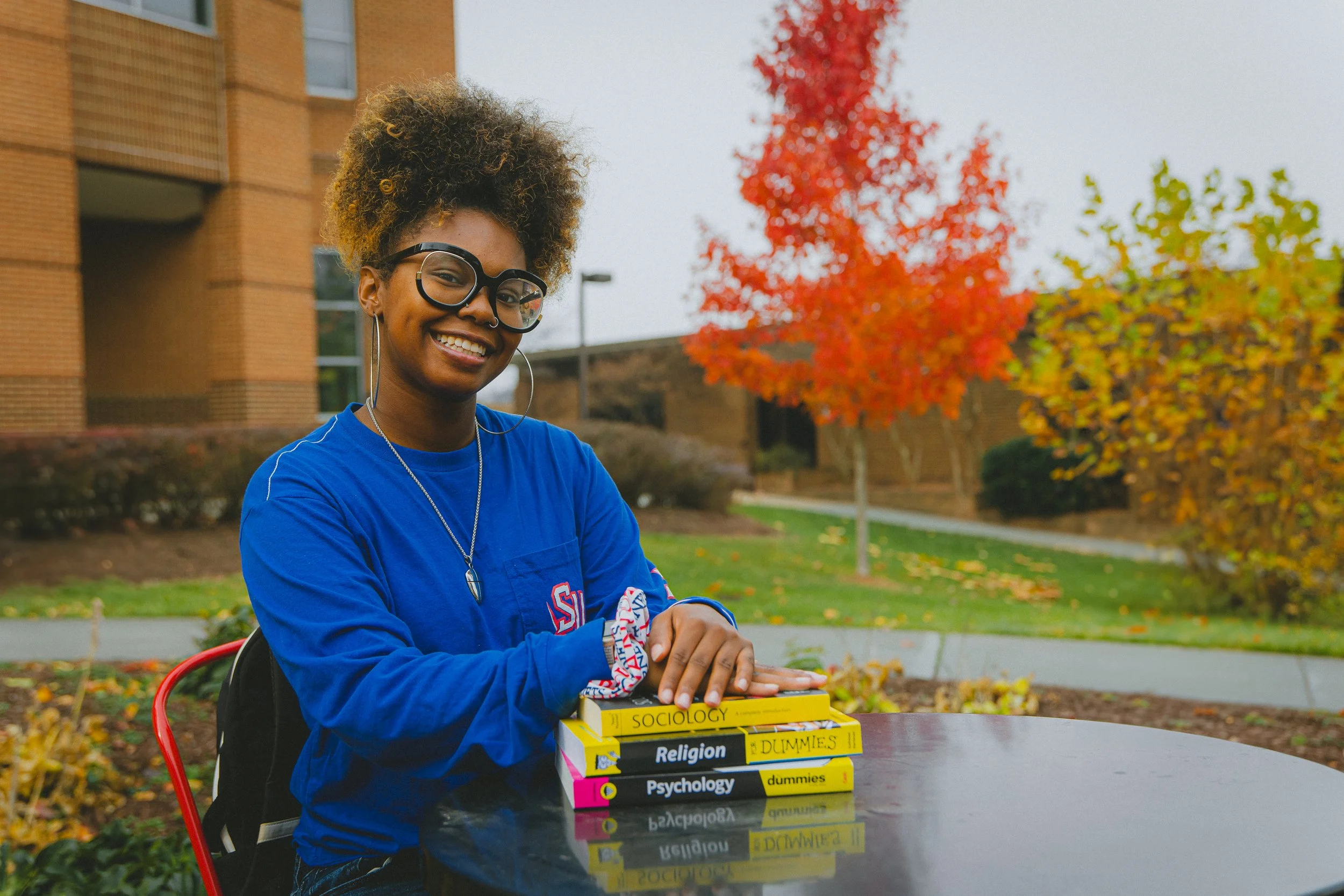 A young woman with glasses and a curly hairstyle, wearing a blue shirt, is sitting outside at a table. She is smiling and resting her hand on a stack of four books about sociology, religion, psychology, and a 'Dummies' guide. The background features 