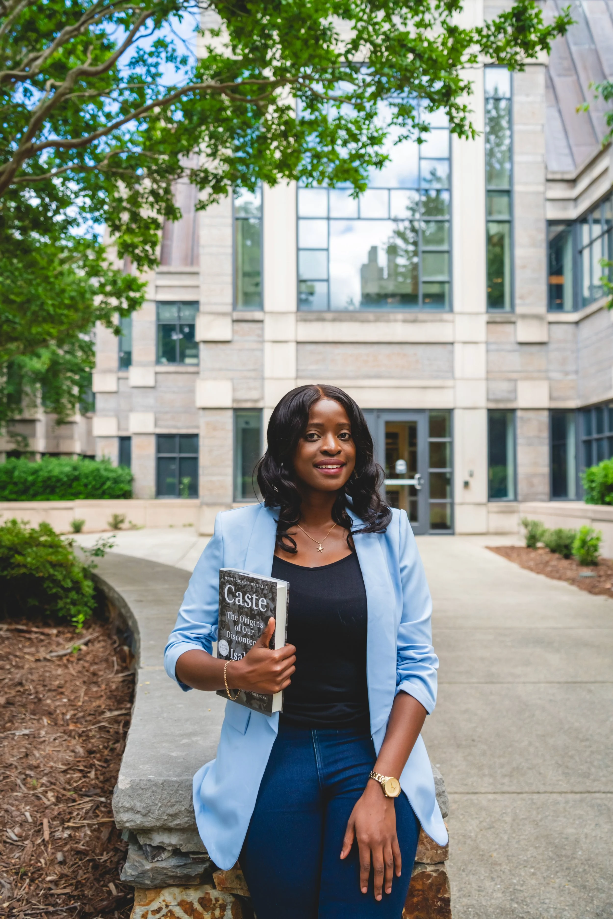 A woman holding a book titled 'Caste: The Origins of Our Discontent' standing outdoors in front of a modern building with large glass windows, surrounded by greenery.