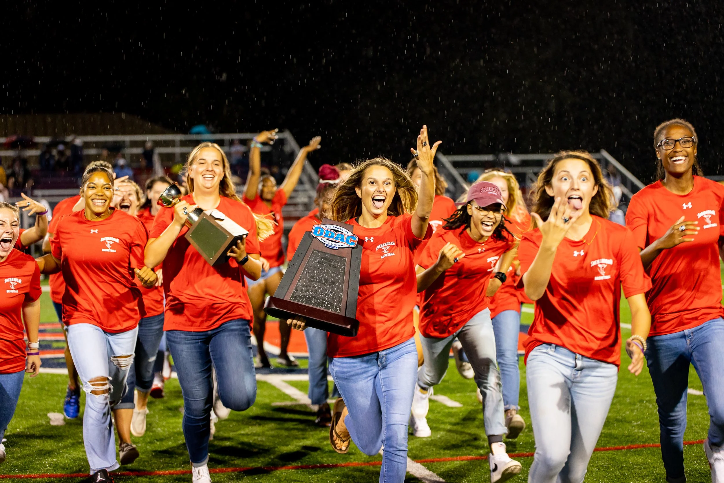 Group of women running and celebrating on football field at night, some holding trophies, wearing red shirts.