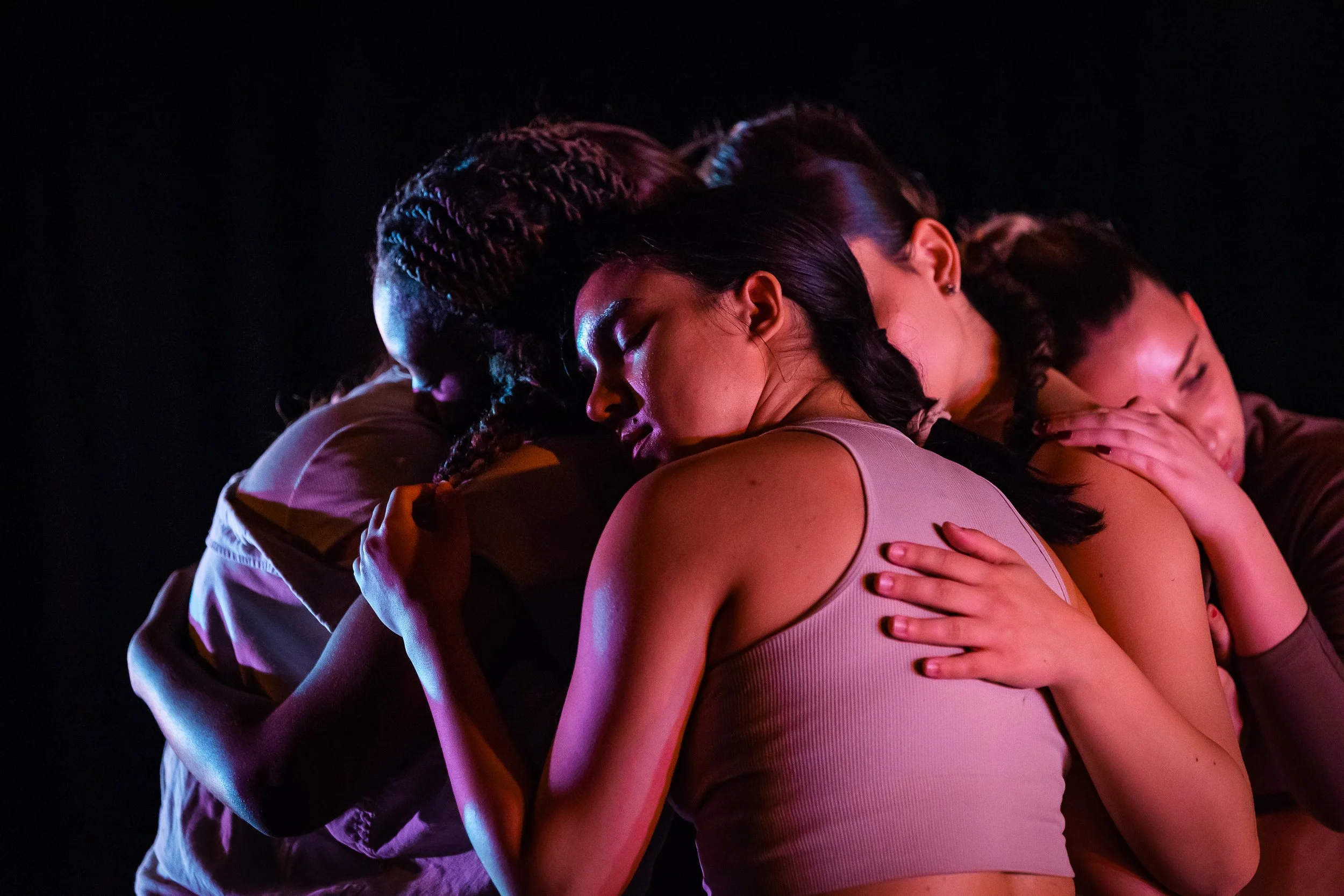 Group of five people embracing in a close hug, with their eyes closed, in a dark setting with dramatic lighting.