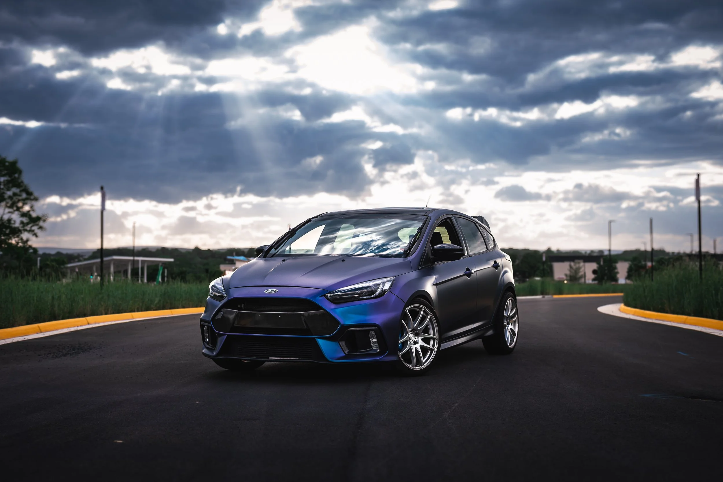 A sleek, black and blue Ford hatchback car parked on a curved asphalt road under a cloudy sky with sun rays breaking through.