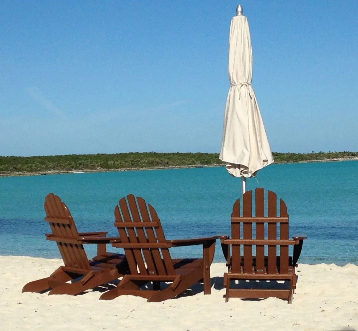 Three empty beach chairs beside a closed, canvas umbrella look out over an aqua blue waterway.
